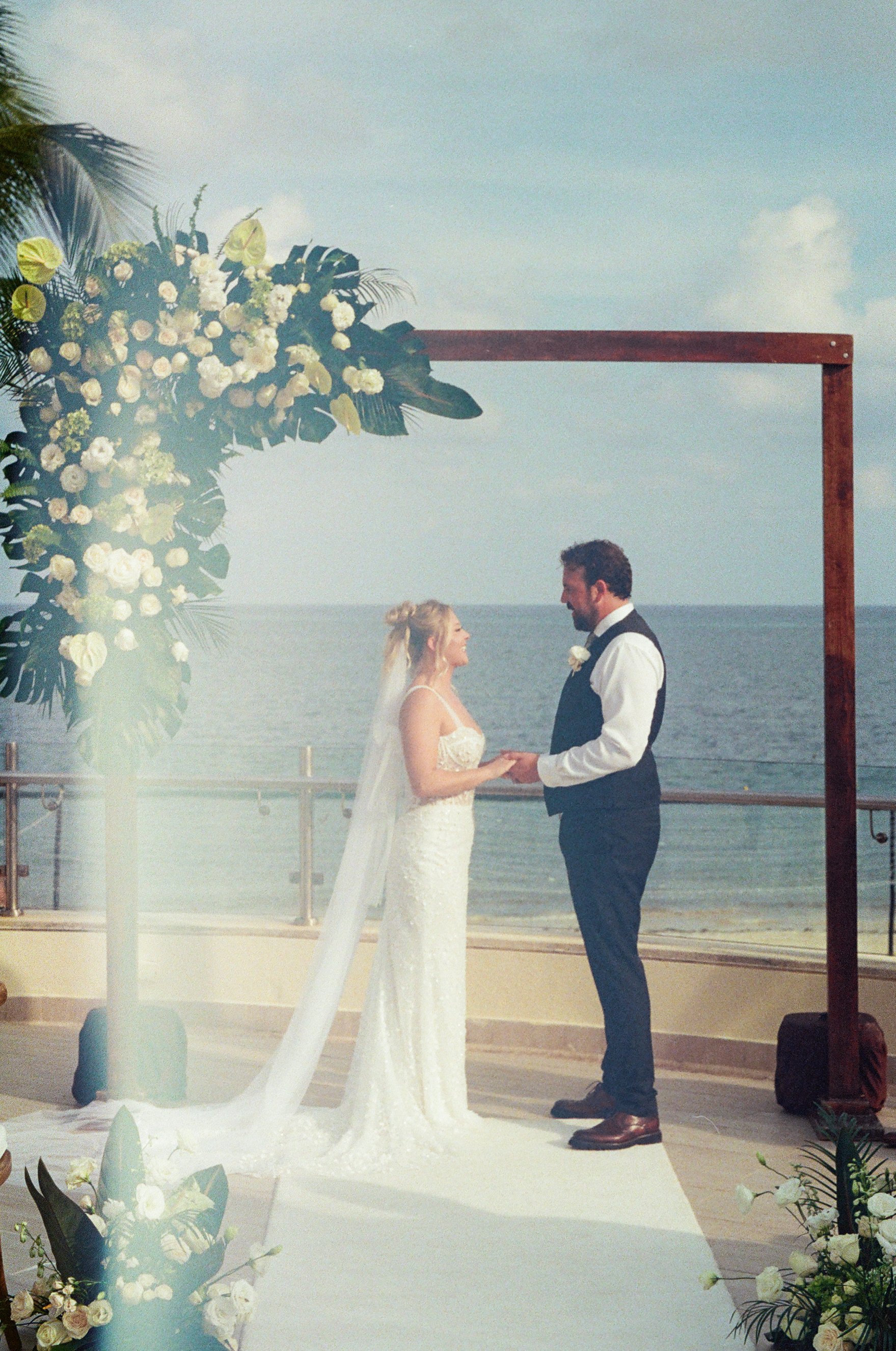 A bride and groom stand holding hands during their wedding ceremony by the ocean, with a floral arch and decorative flowers around.