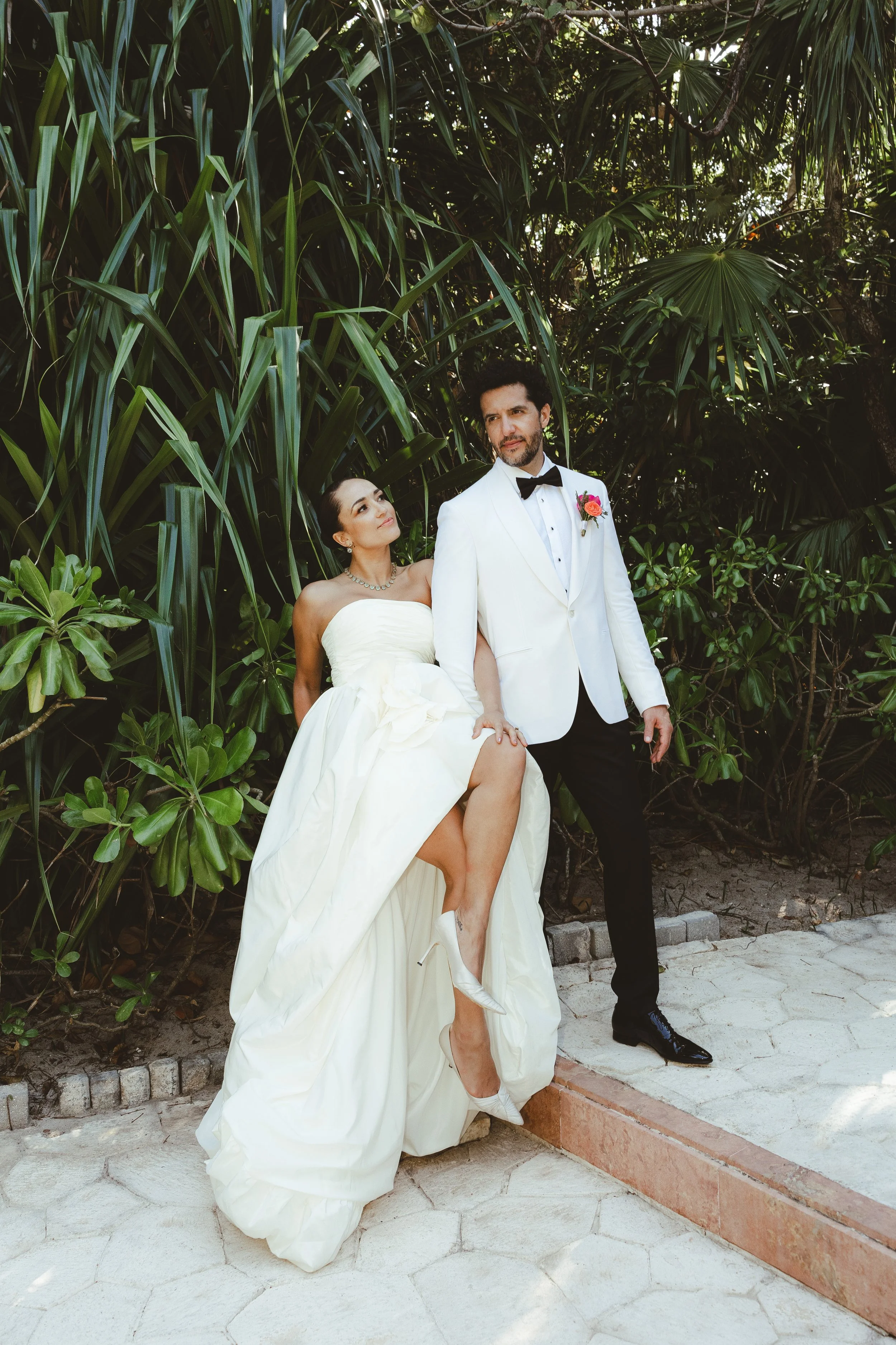 A bride in a strapless white wedding gown and white heels, and a groom in a white tuxedo jacket with black pants and a bow tie, standing on a paved outdoor area surrounded by dense green tropical foliage.