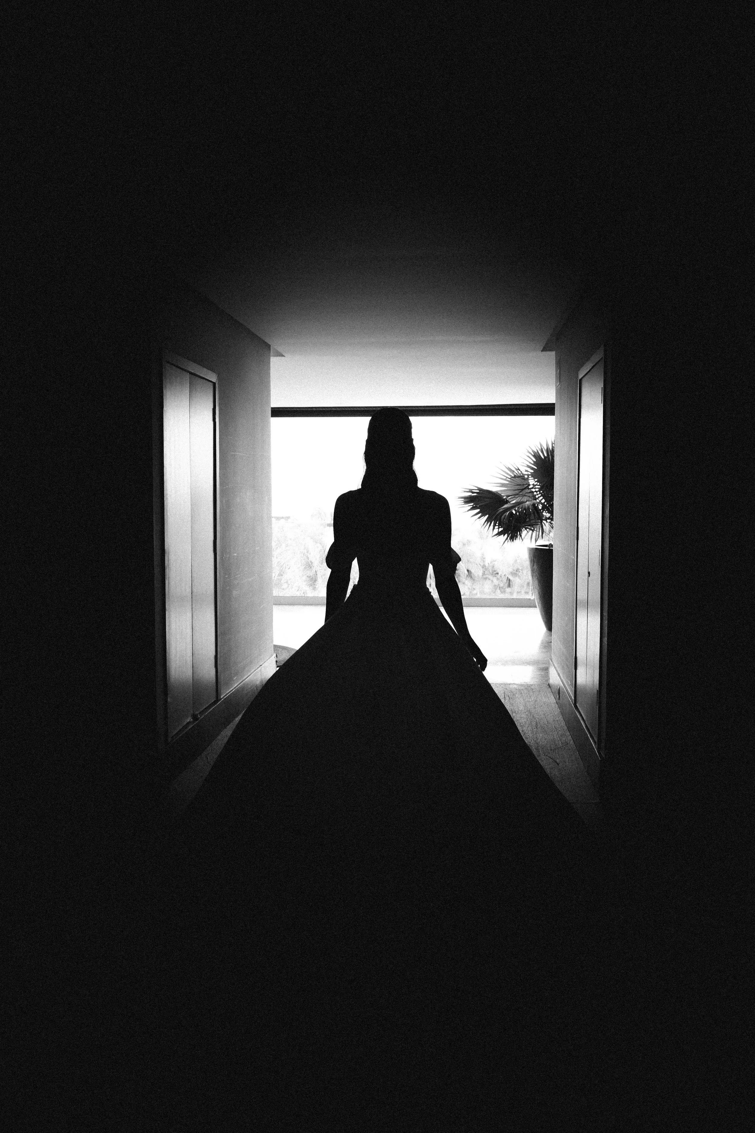 A bride walking towards a balcony with natural light and a potted plant beside them. Black and white artistic wedding photography.