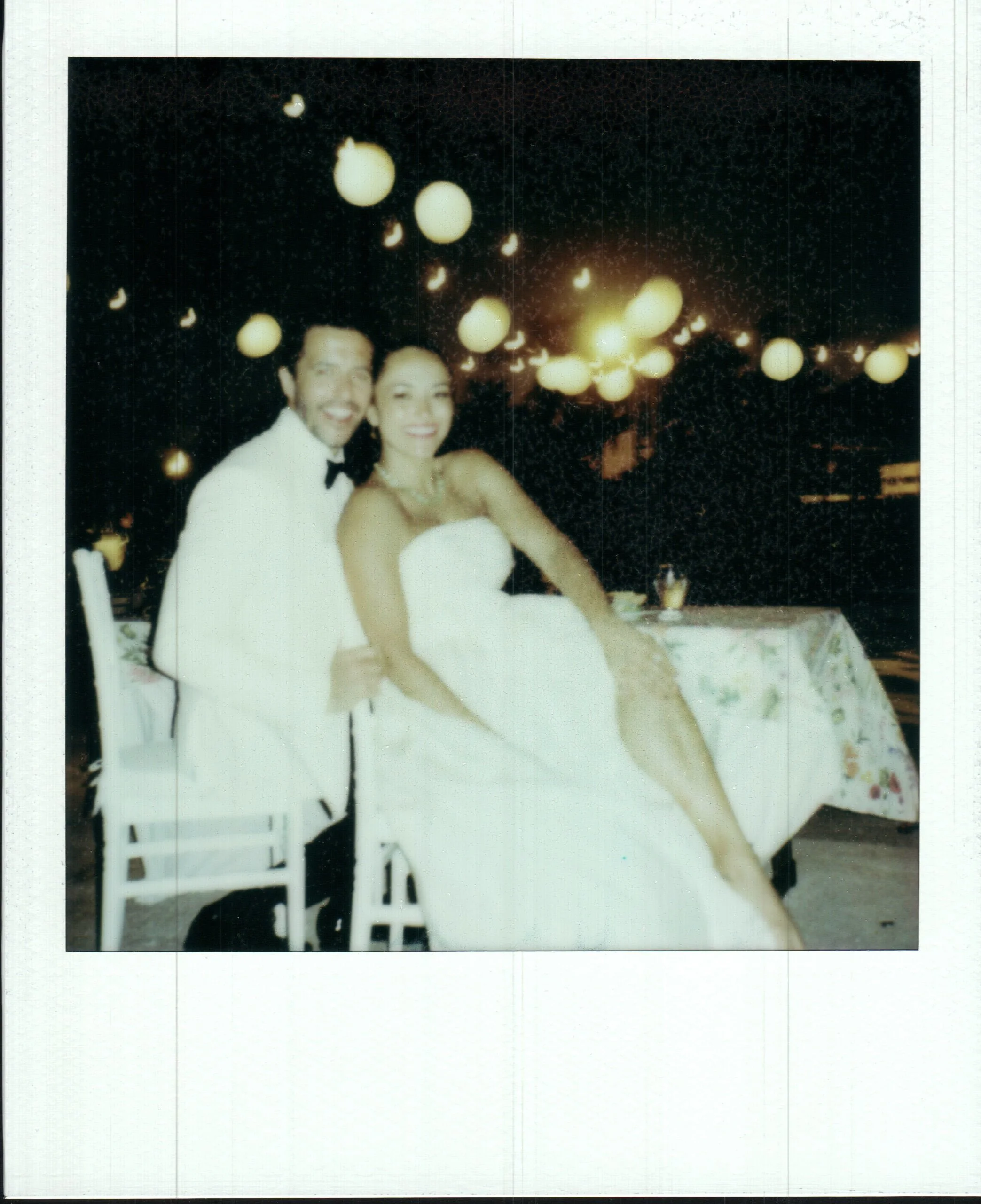 A man and woman dressed as a groom and bride in wedding attire, sitting at a table with string lights hanging in the background during a night celebration.