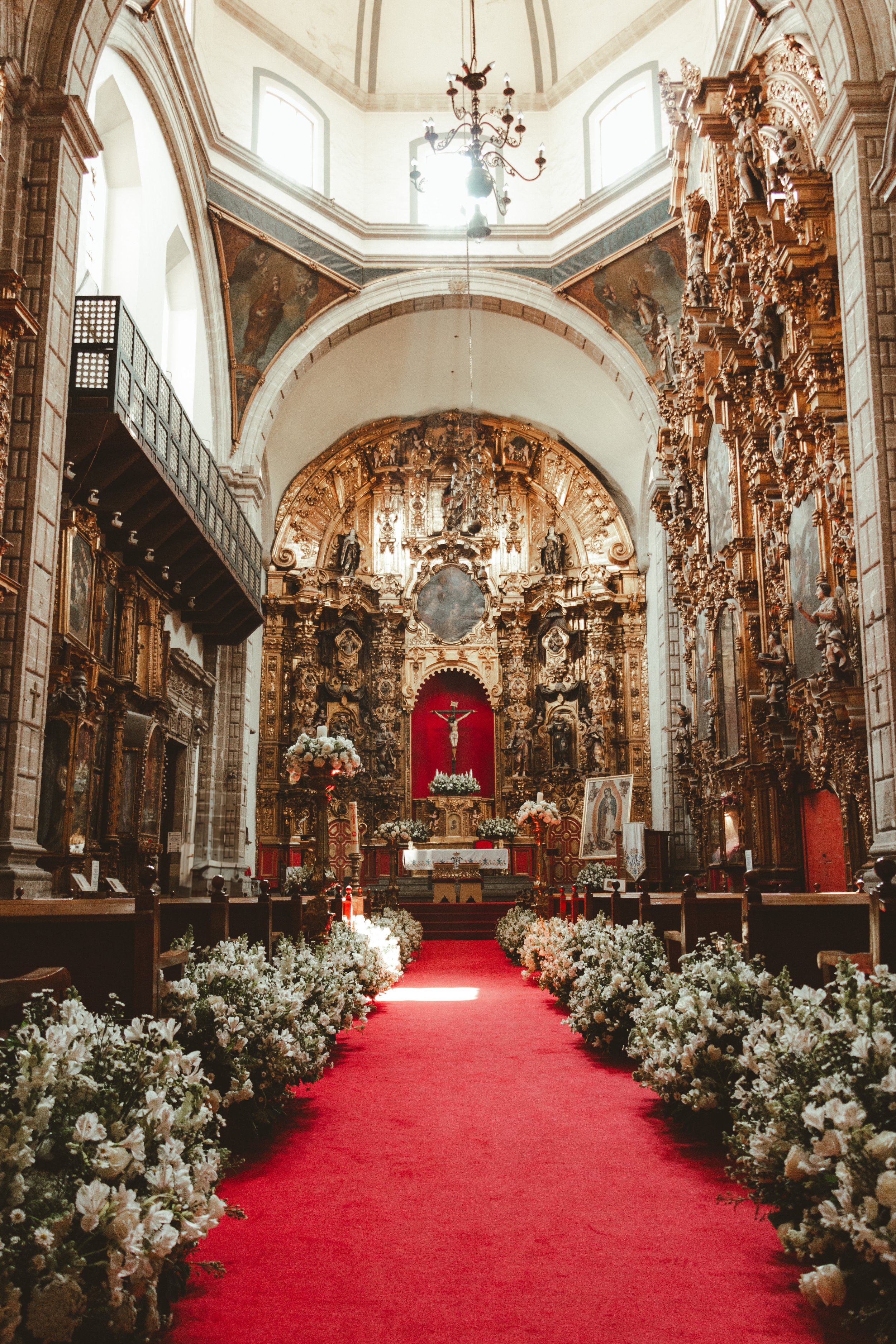 Interior of a church in Ciudad de Mexico decorated with flowers for a wedding, featuring a red carpet aisle, ornate gold altar, and floral arrangements along the aisle.