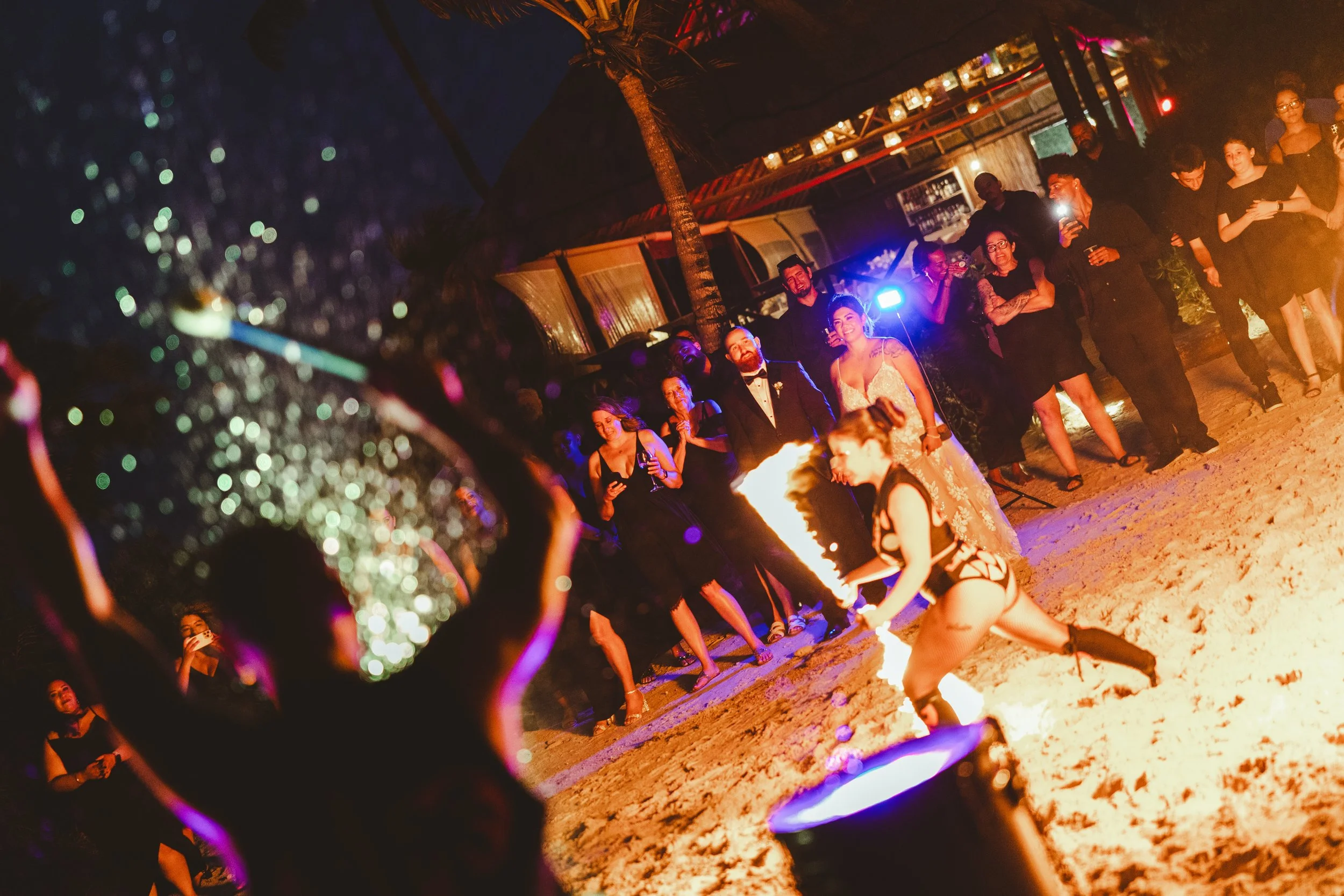 A woman performing a fire dance on a sandy beach at night, with an audience watching and taking photos, illuminated by colorful stage lighting.