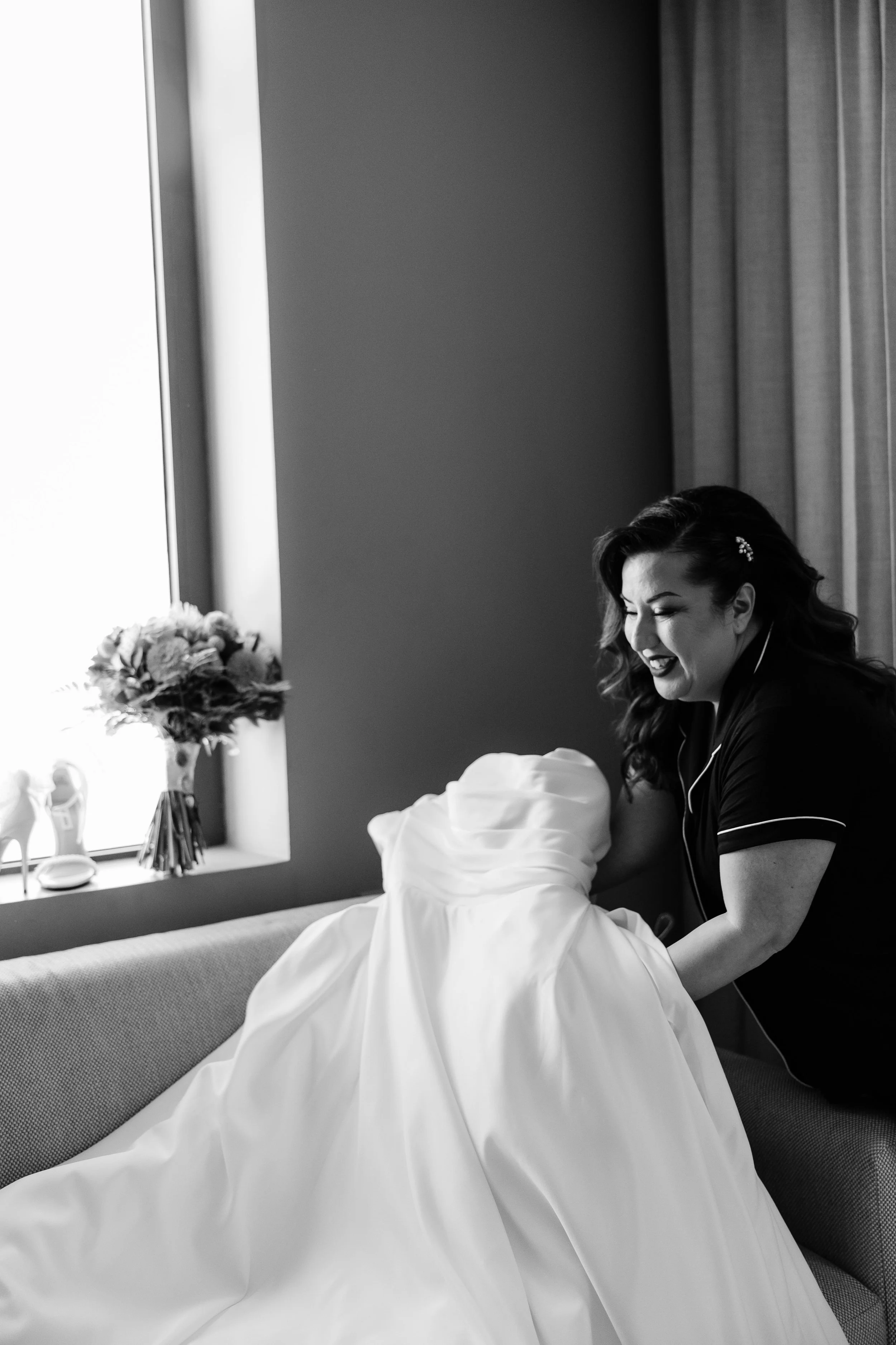 A woman smiling and wearing earphones, holding a wedding dress, sitting on a sofa near a window with a bouquet of flowers nearby.