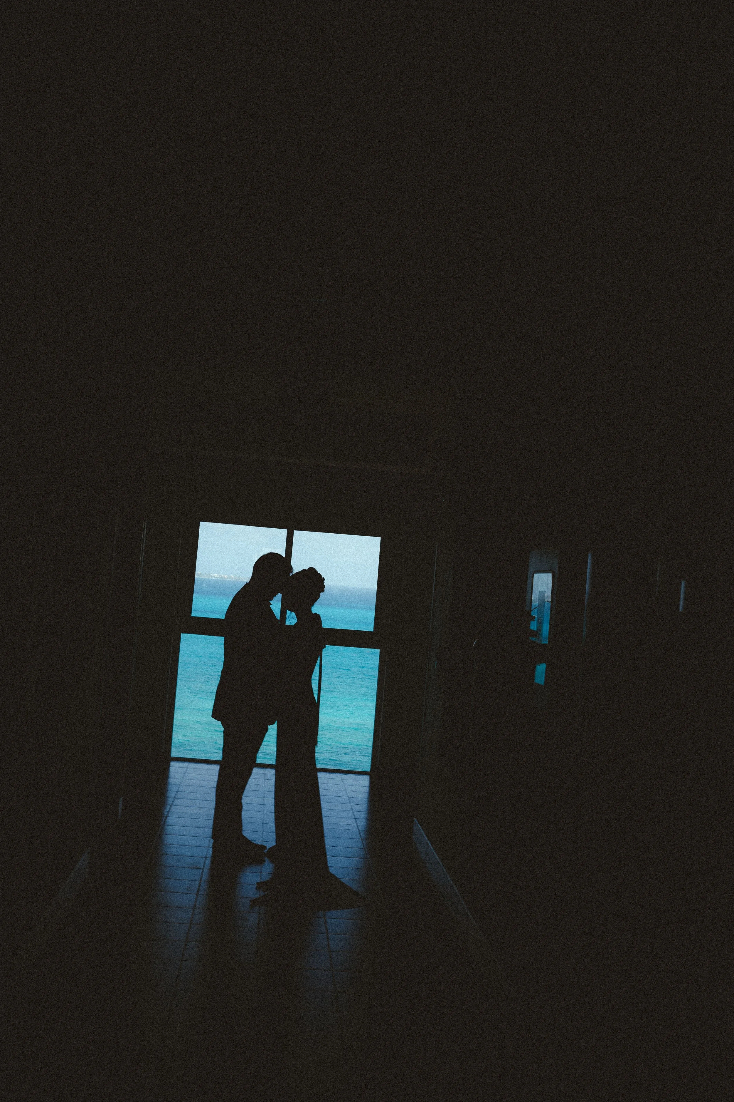 Silhouetted couple standing close together at a window, with ocean view in the background, during their wedding in cancun.