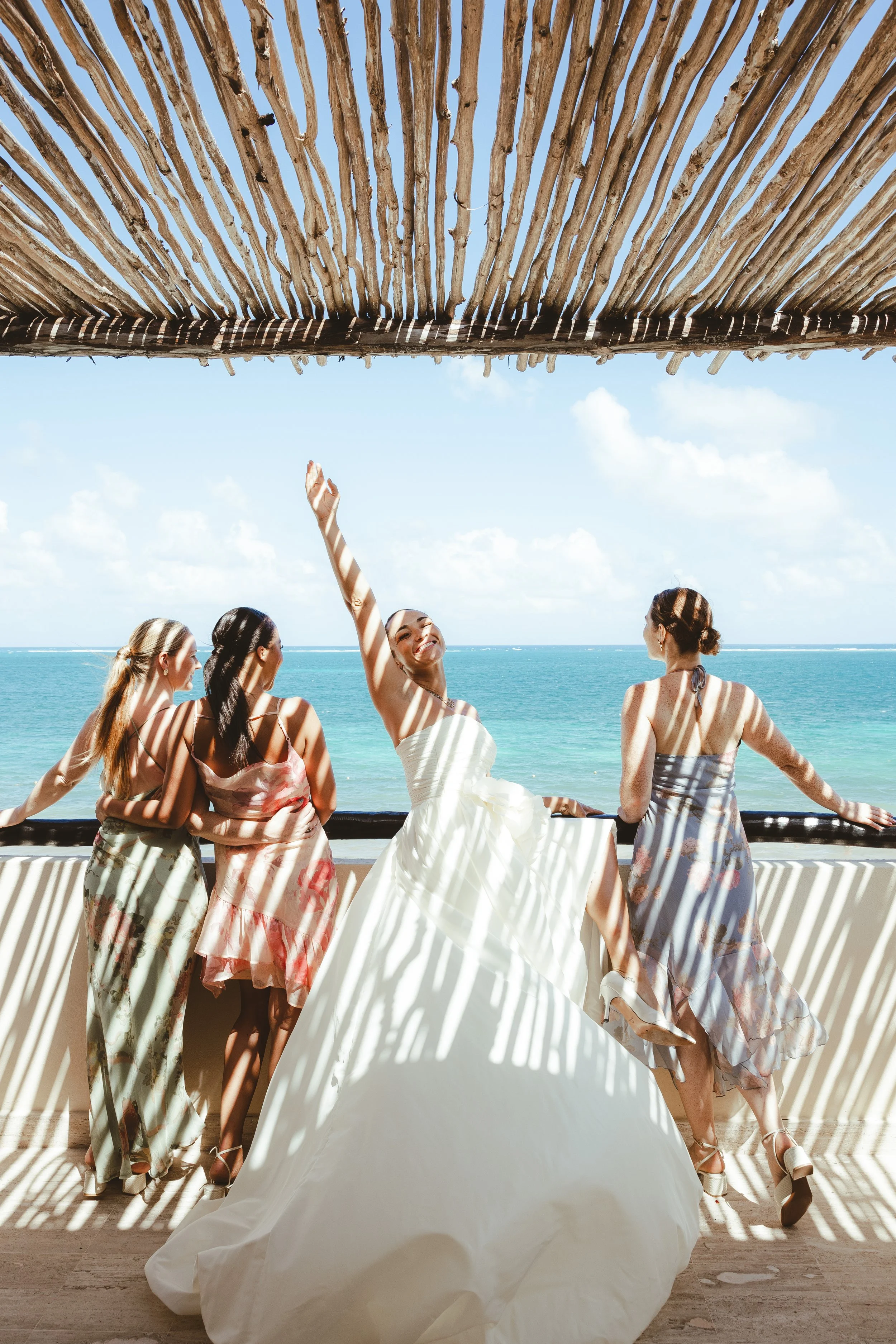 A bride and her four bridesmaids enjoying a sunny day at the beach, standing on a balcony with ocean view, casting shadows through a wooden pergola above.