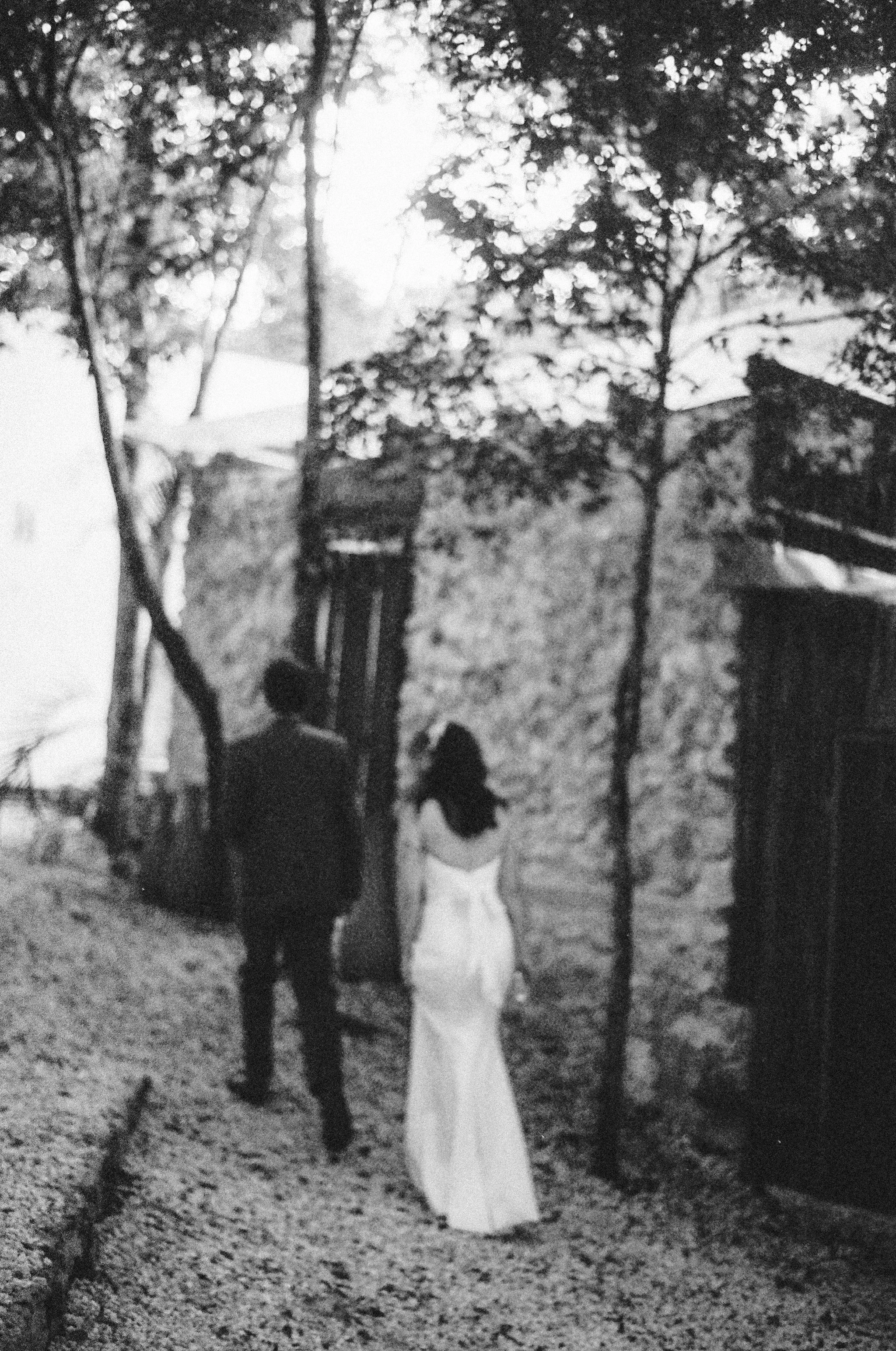 A black and white film photo of a bride and a groom walking away together down a gravel path, surrounded by trees and old stone walls, with the woman wearing a long white dress.