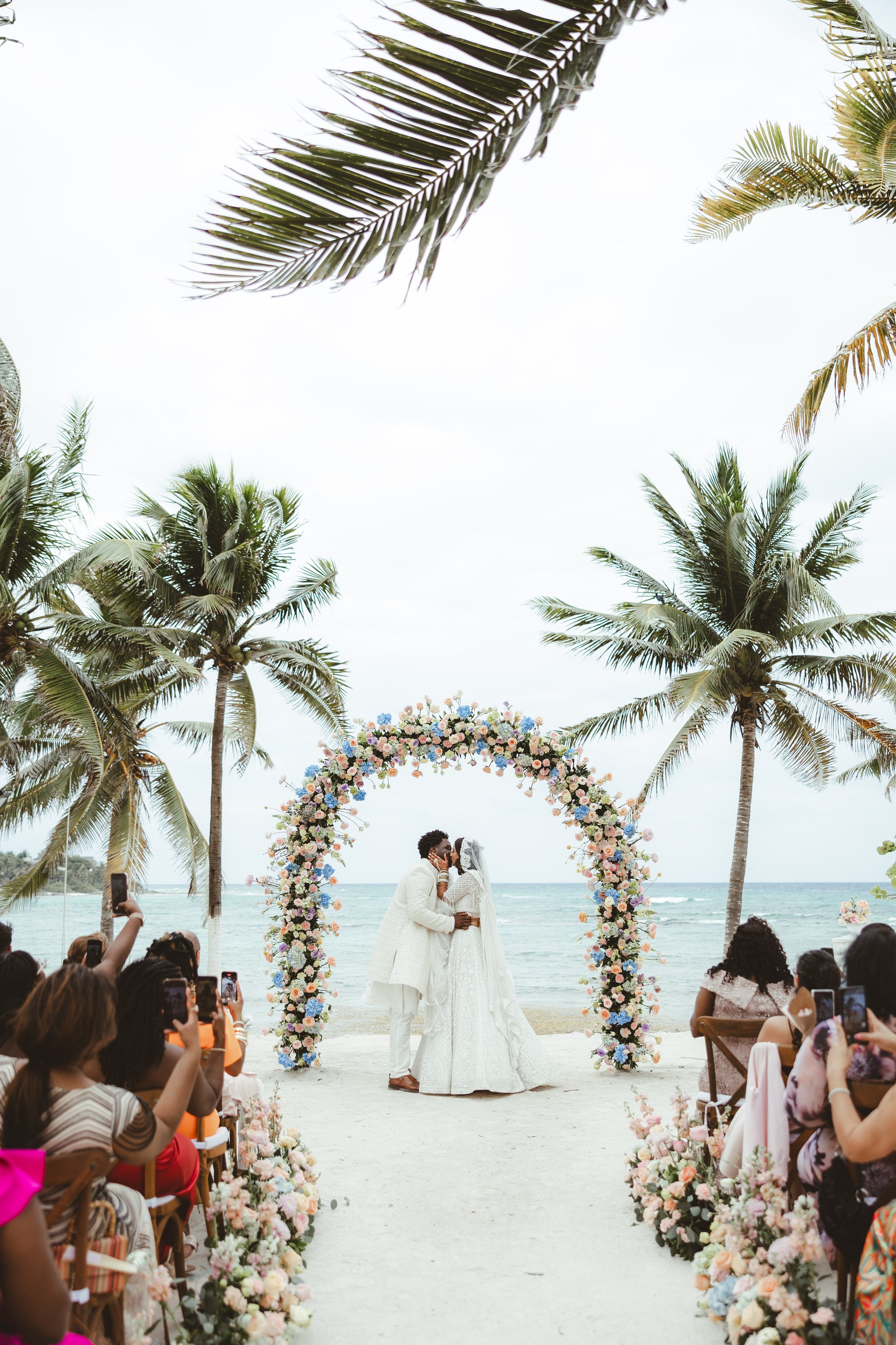 A couple in wedding attire sharing a kiss under a floral arch on a beach during a wedding ceremony, with guests taking photos and palm trees surrounding the scene. Blue Venado.