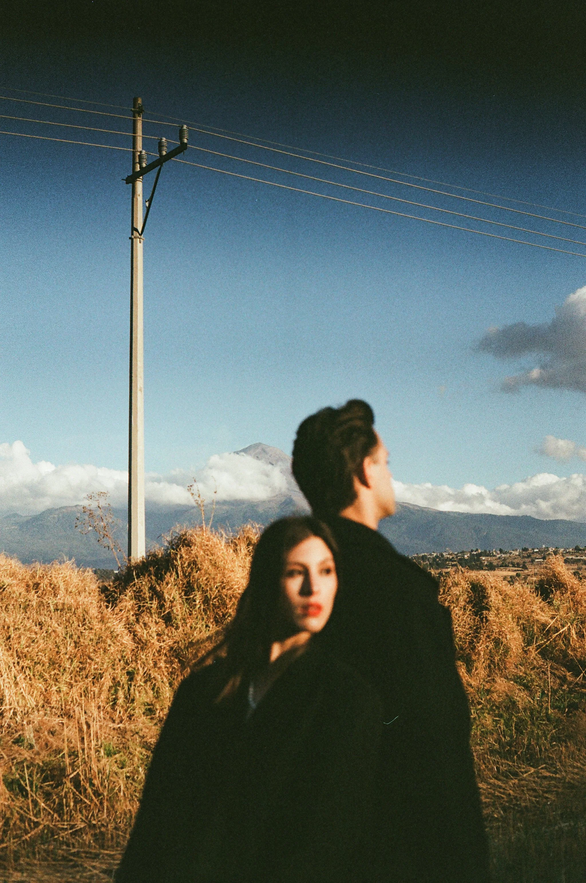 A couple standing in a field of tall grass with mountains and a power line in the background. The woman has long dark hair and is looking at the camera; the man has short dark hair and is looking away.