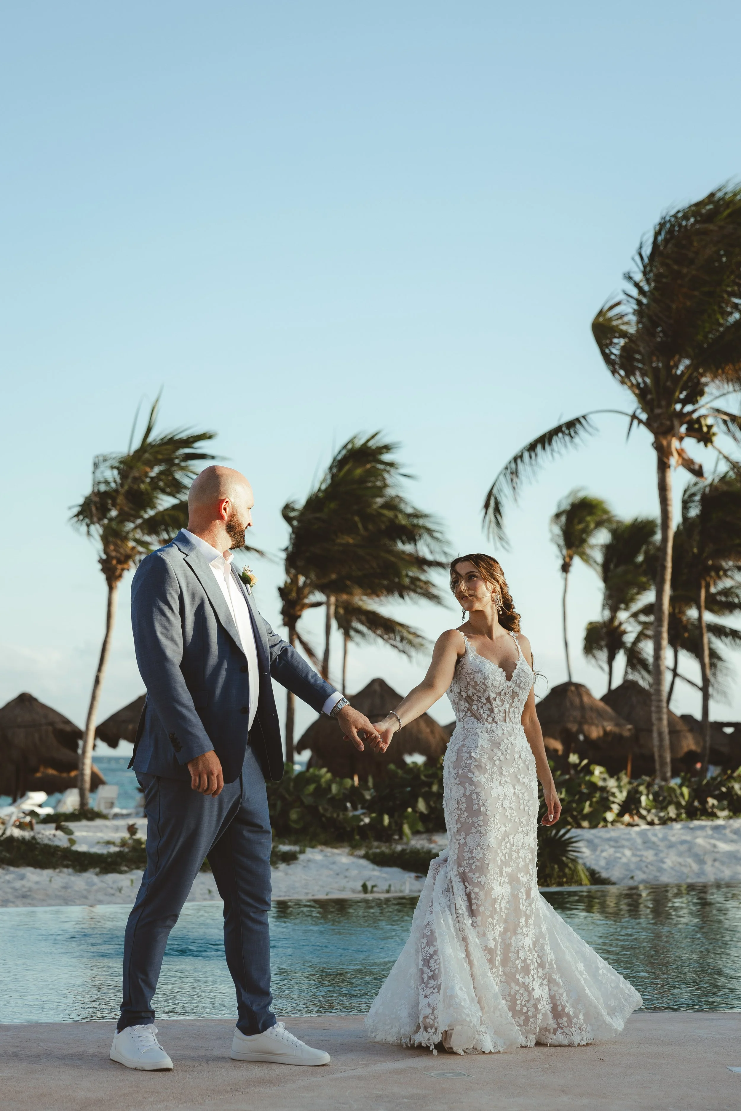 A bride and groom holding hands during their wedding ceremony on a beach with palm trees and thatched umbrellas in the background.