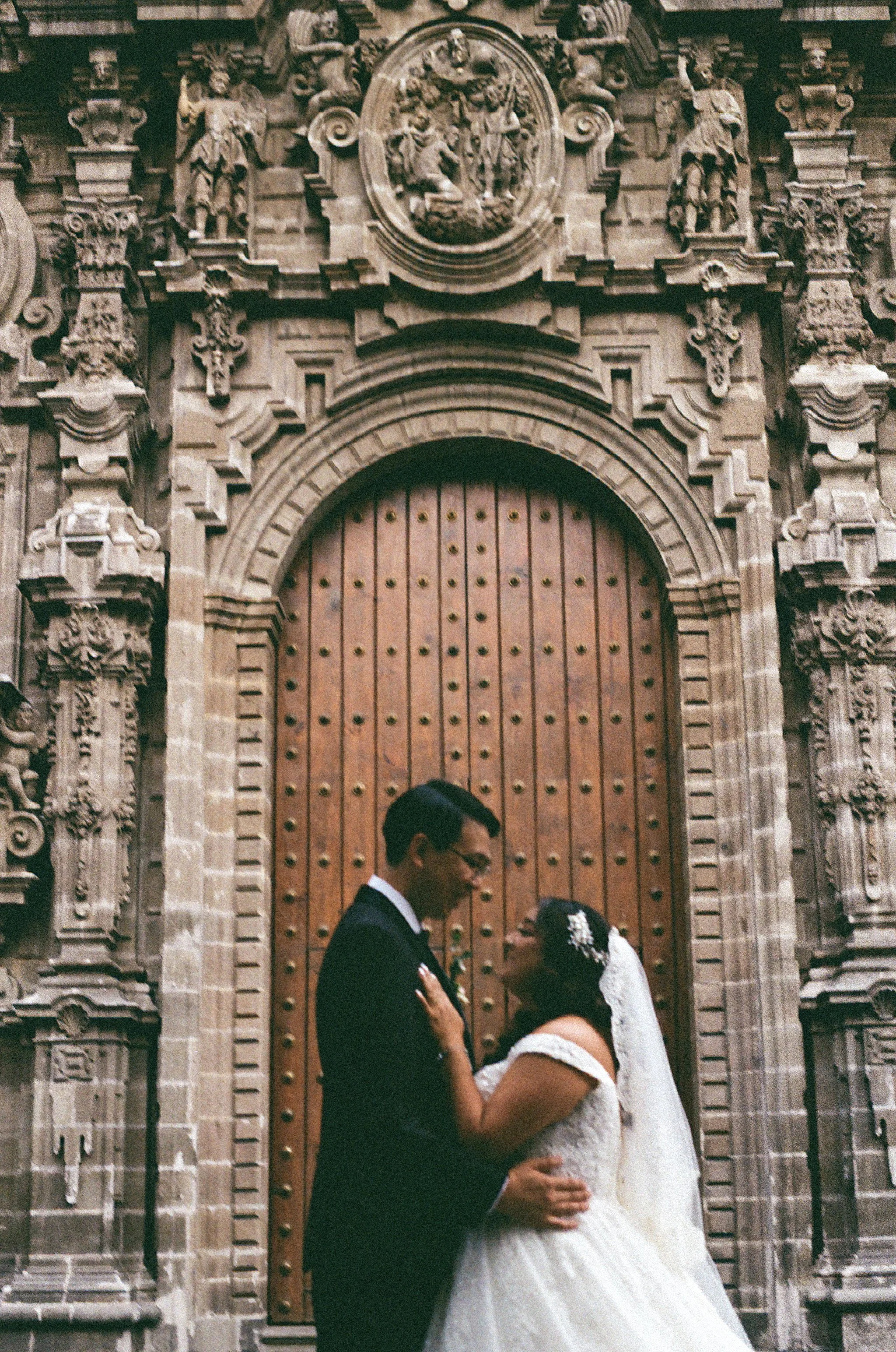 A bride and groom standing close together in front of an ornate, carved stone door, sharing a moment on their wedding day.