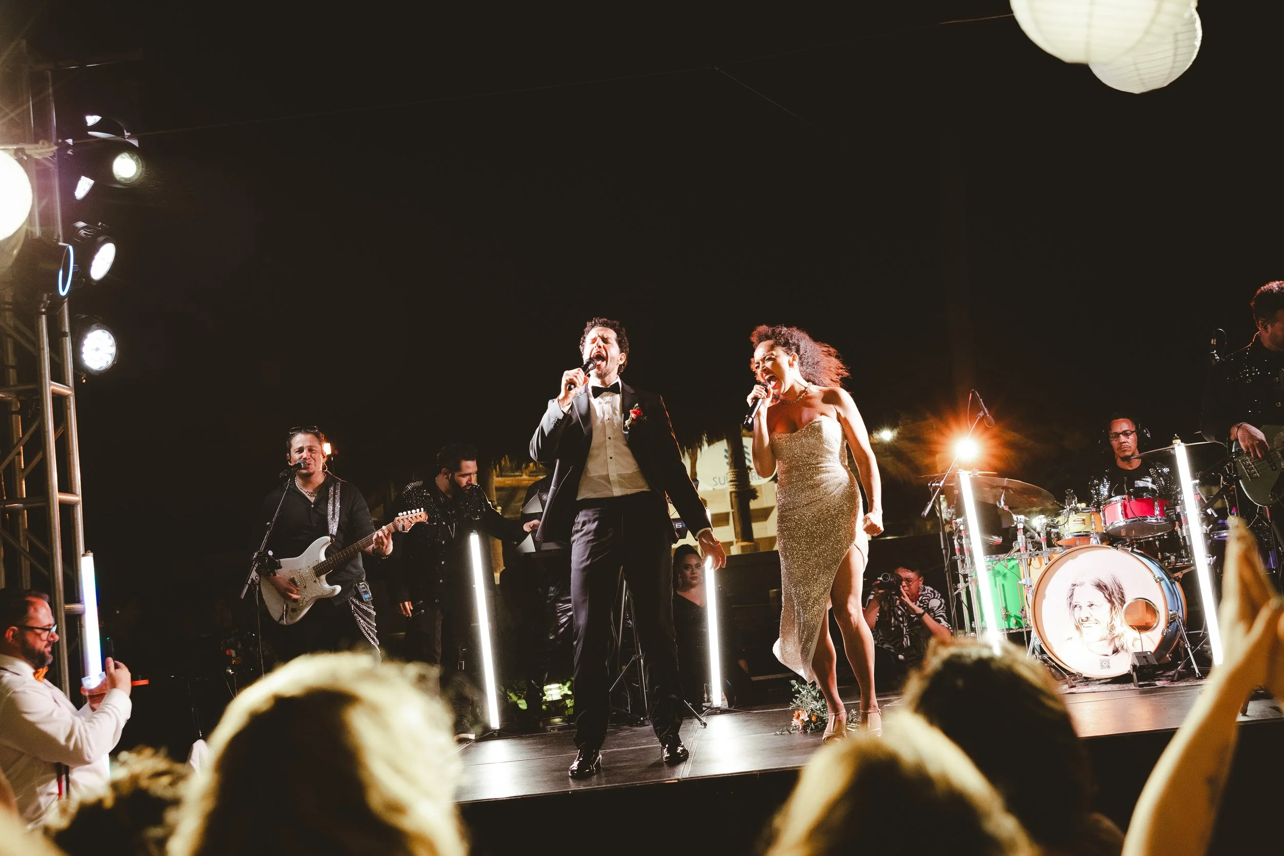 A live music performance at night featuring a male singer and a female singer on stage, with a band playing instruments in the background, audience members in the foreground, and bright stage lights.