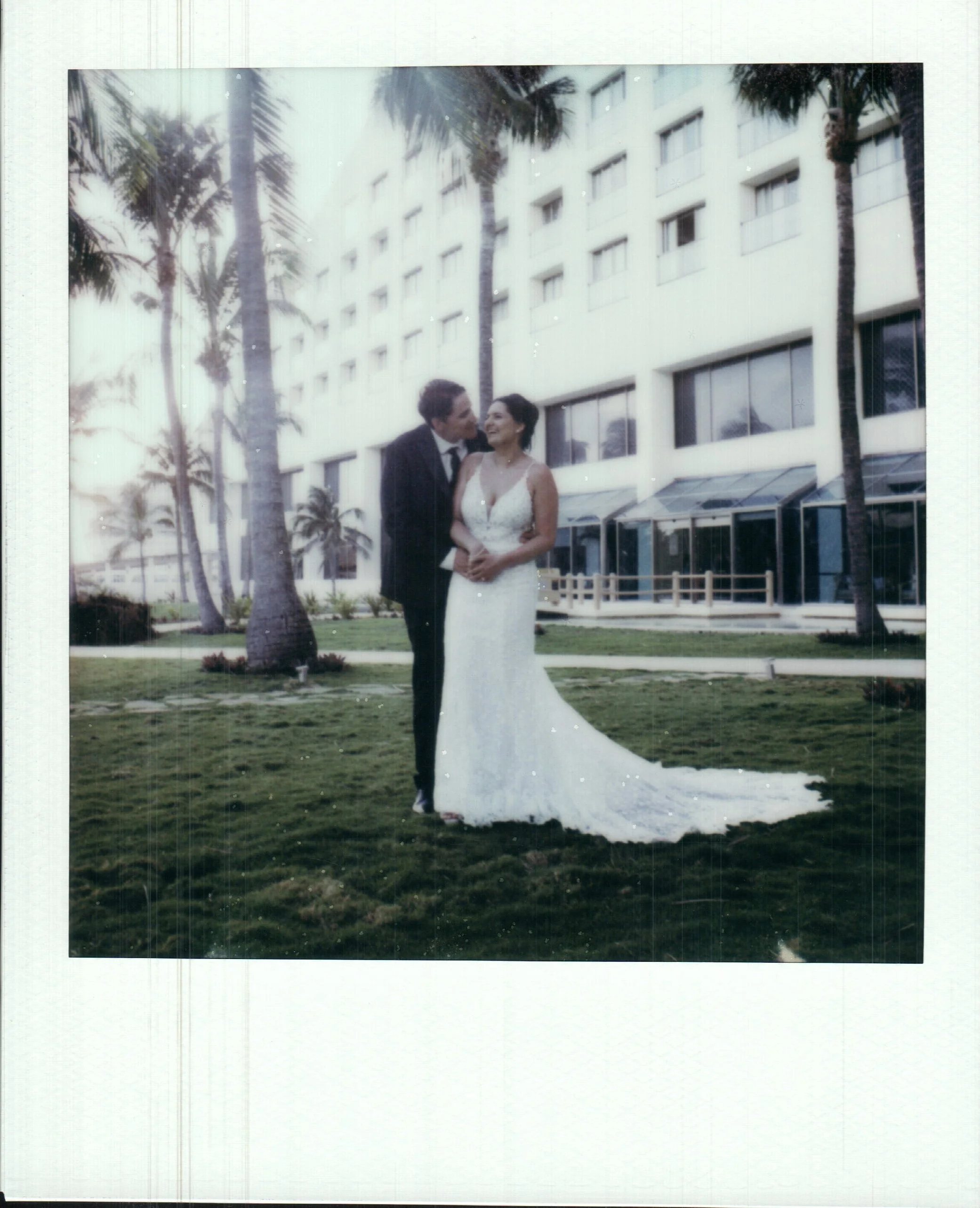 A bride and groom standing on grass in front of a white building with palm trees. The bride is wearing a white wedding gown, and the groom is in a black suit.
