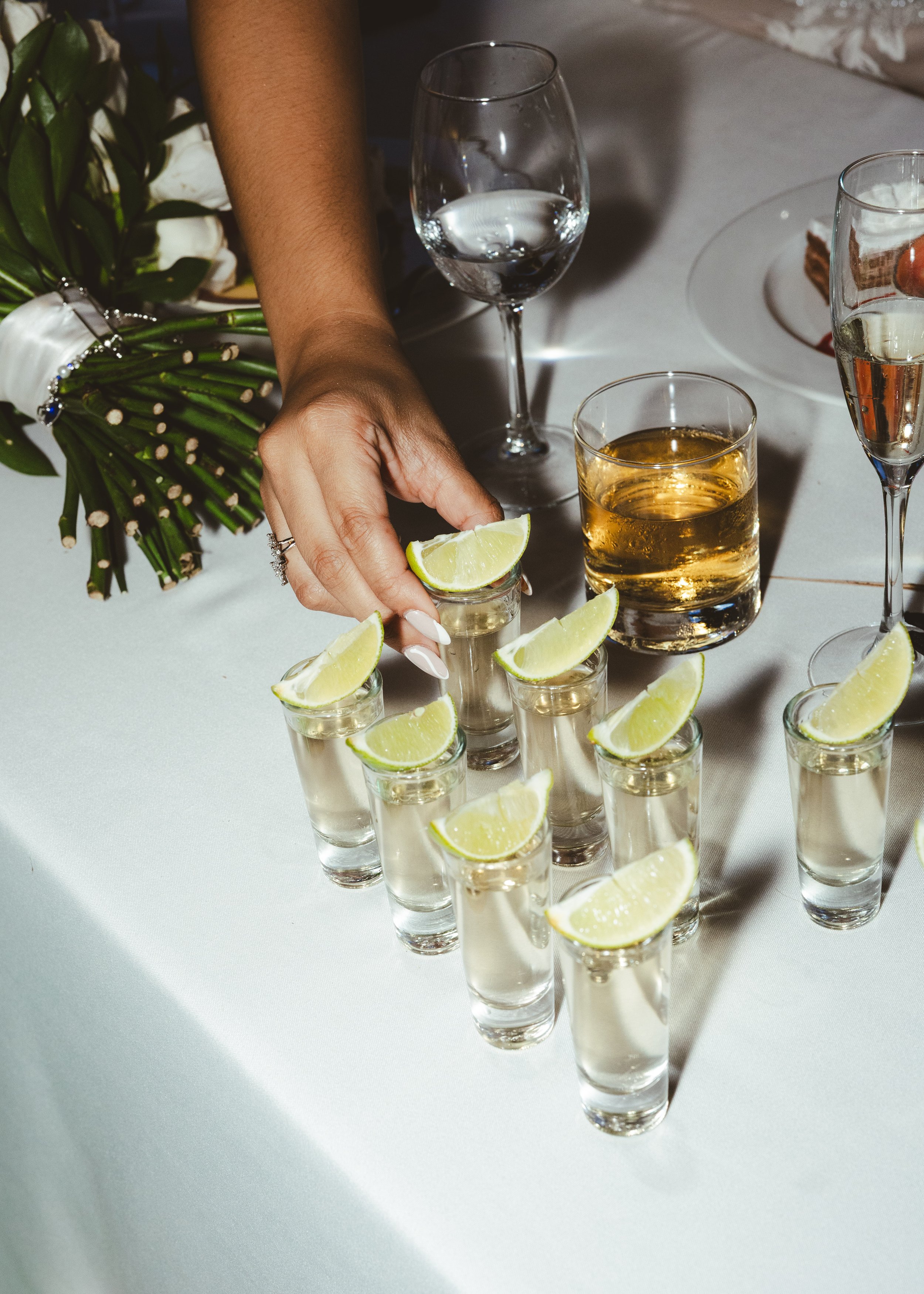 A person arranging shot glasses filled with clear liquid, each garnished with a lemon wedge, on a white table. There are glasses of water, whiskey, and champagne, along with a bouquet of white flowers.