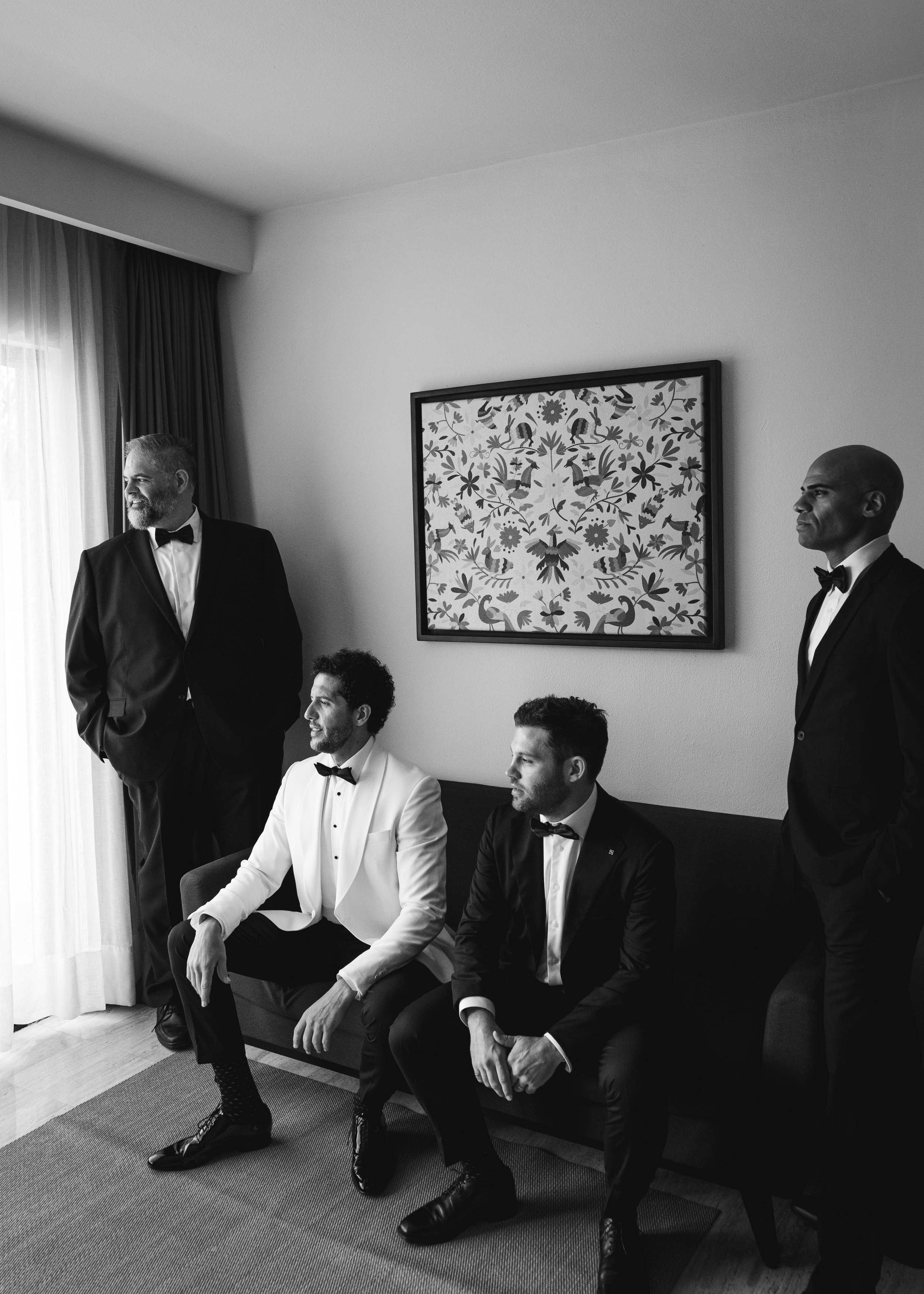 Four men in tuxedos with bowties in a hotel room, two seated on a sofa and two standing, all looking in different directions, in front of a window with curtains and a framed floral artwork on the wall, in black and white.