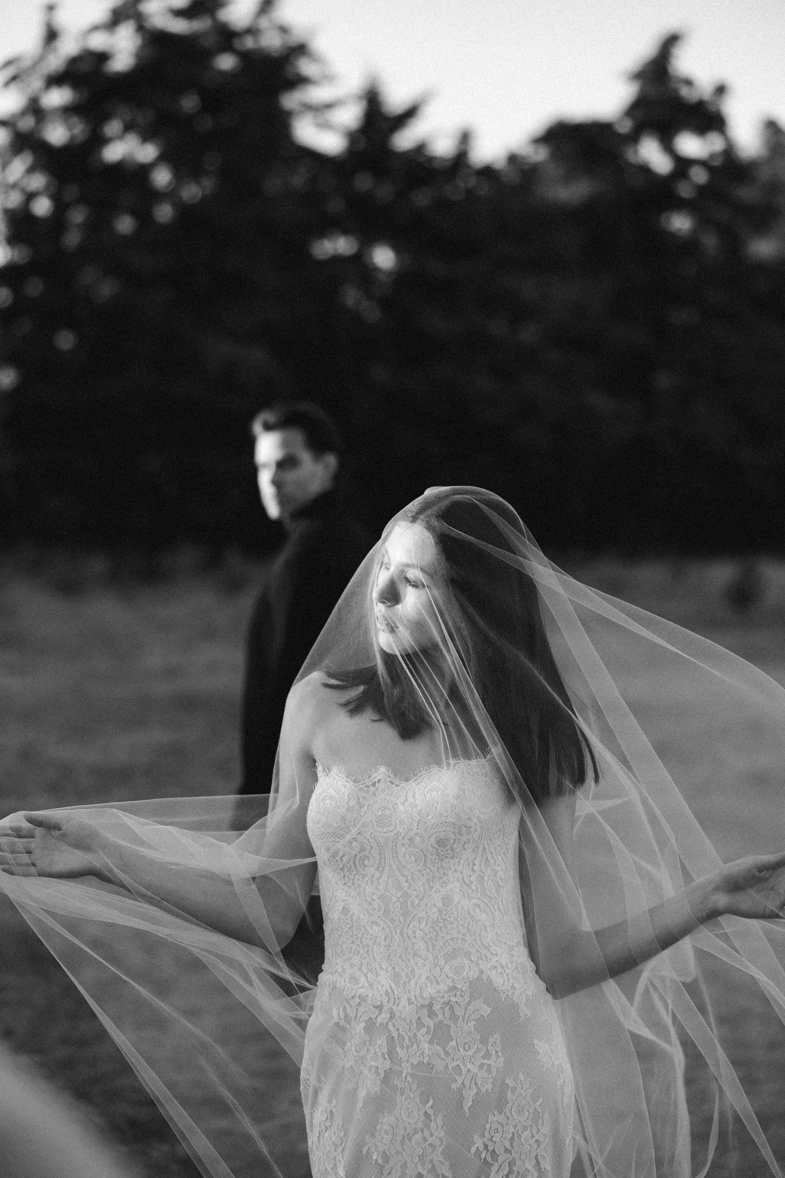 A bride in a strapless lace wedding dress with a veil over her face, outdoors. A man stands in the background among trees, out of focus. Wedding in the woods inspo.