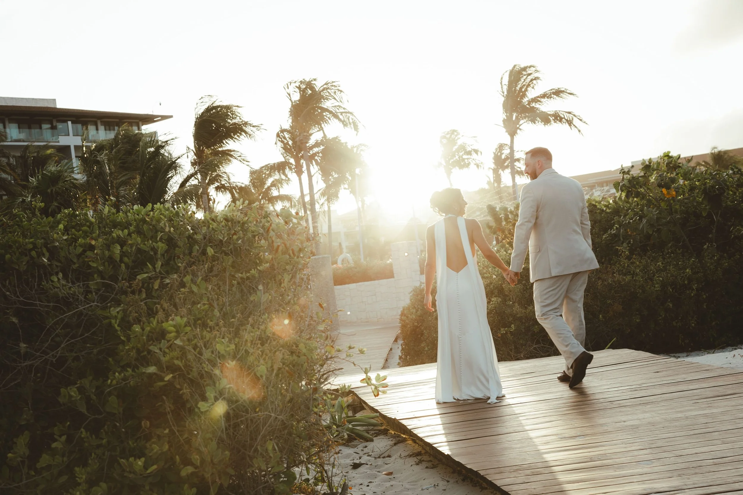 A couple holding hands and walking on a wooden pathway outdoors during sunset, surrounded by green bushes and palm trees. Wedding sunset photography.