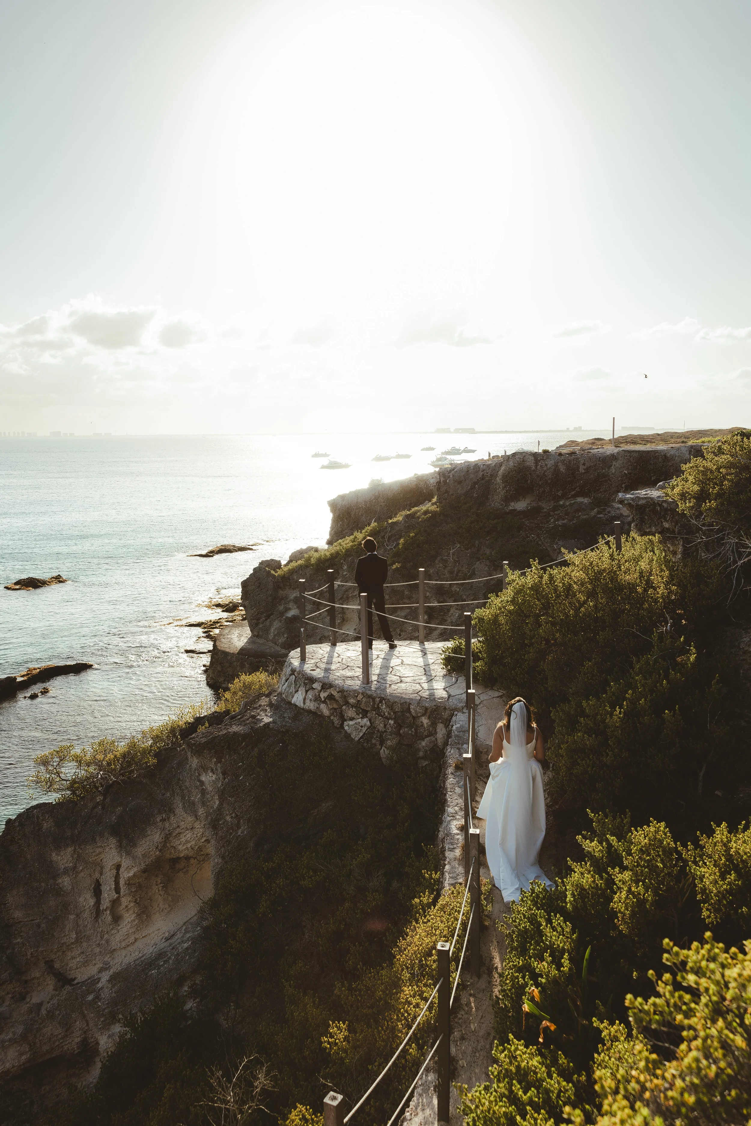 A bride walking along a narrow path on a rocky cliff overlooking the ocean, with a groom standing on a viewing platform nearby, during sunset.