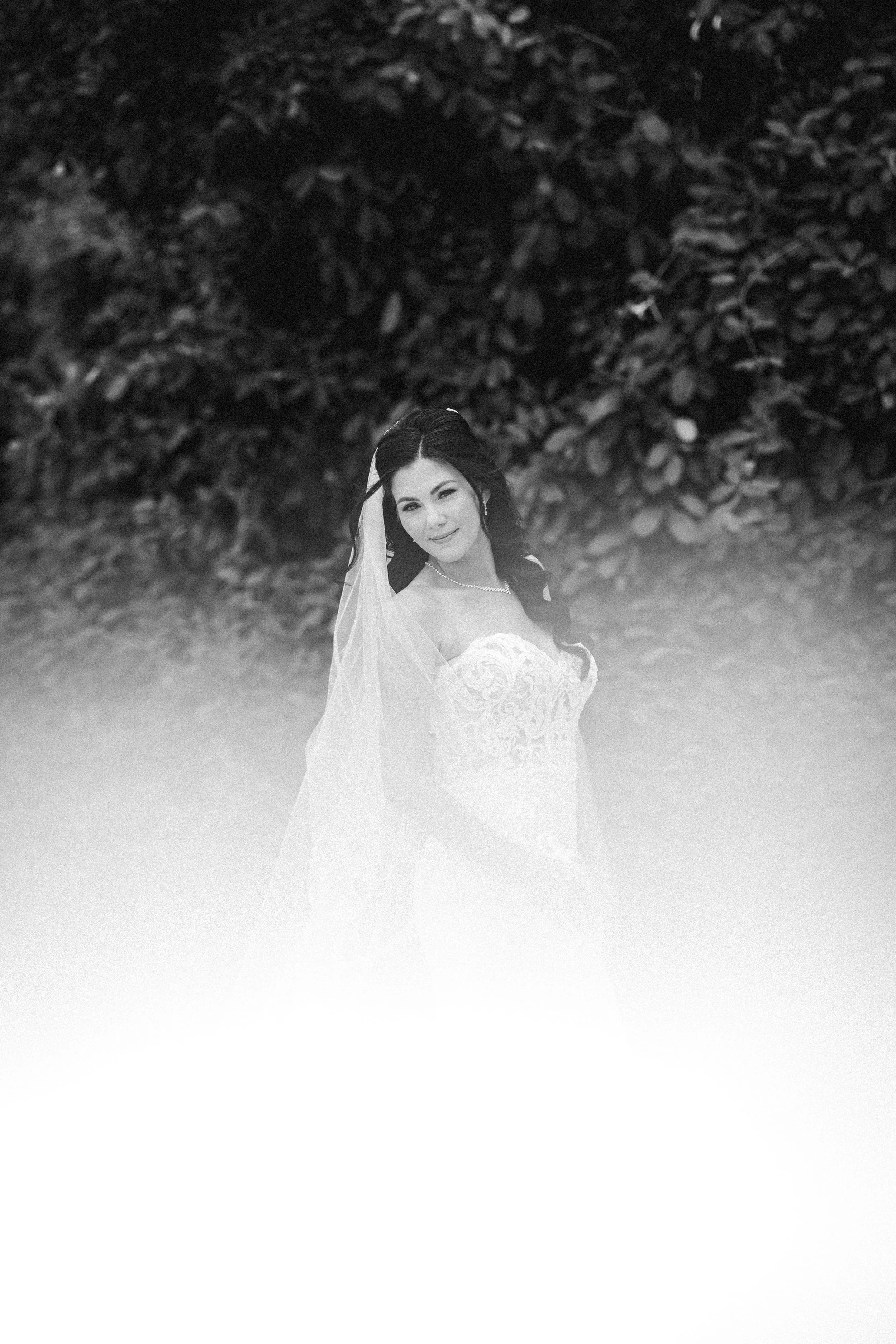 Black and white photo of a bride in a wedding dress with veil, standing outdoors in front of a bush in Paradisus Playa del Carmen.