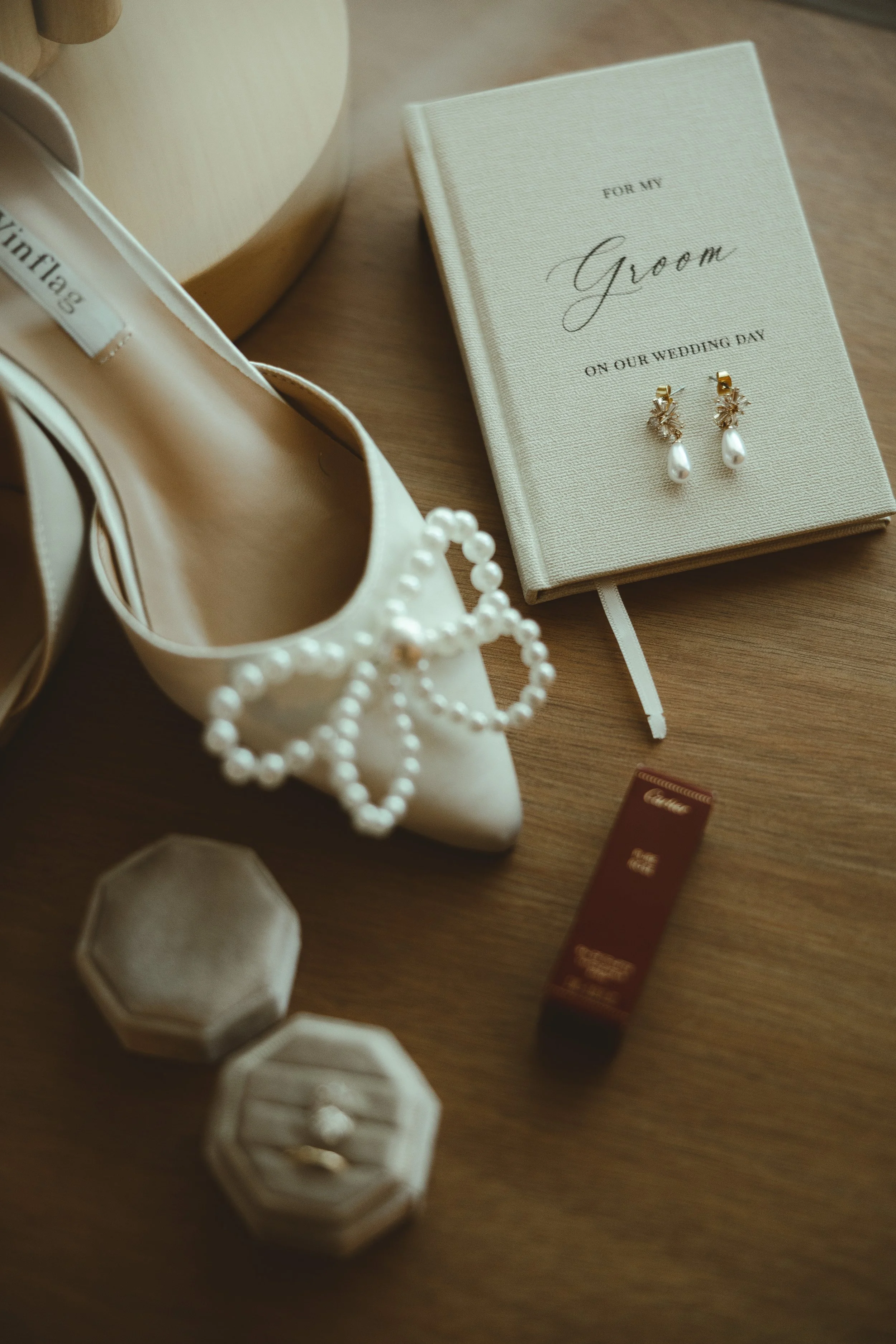 A pair of white wedding shoes, pearl jewelry, a wedding guest book, and a box of lipstick on a wooden surface, prepared for a wedding.