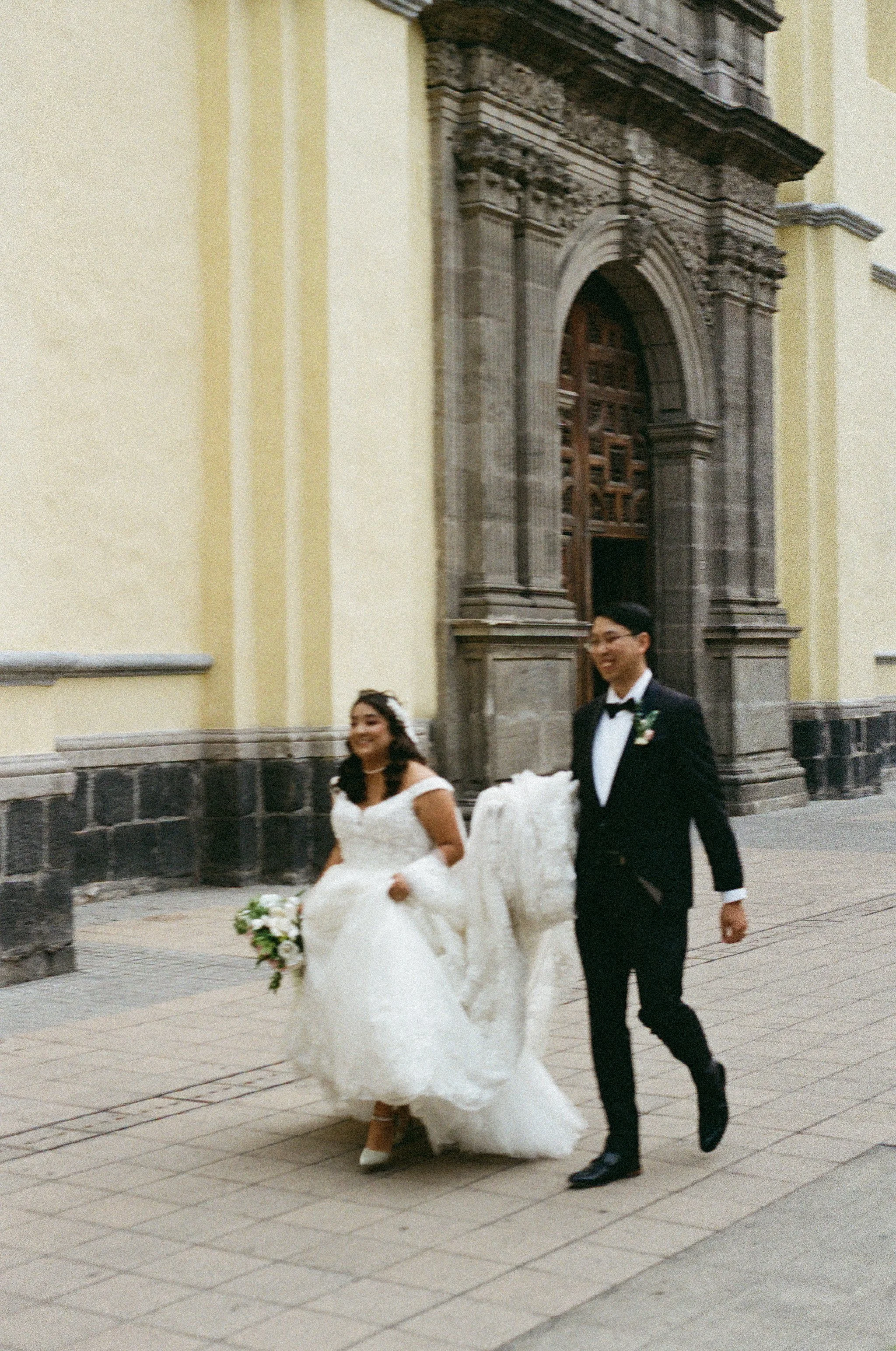A bride and groom walking outside a church, dressed in wedding attire. The bride is wearing a white wedding gown, holding a bouquet of flowers, and smiling. The groom is in a black tuxedo with a bow tie, also smiling.
