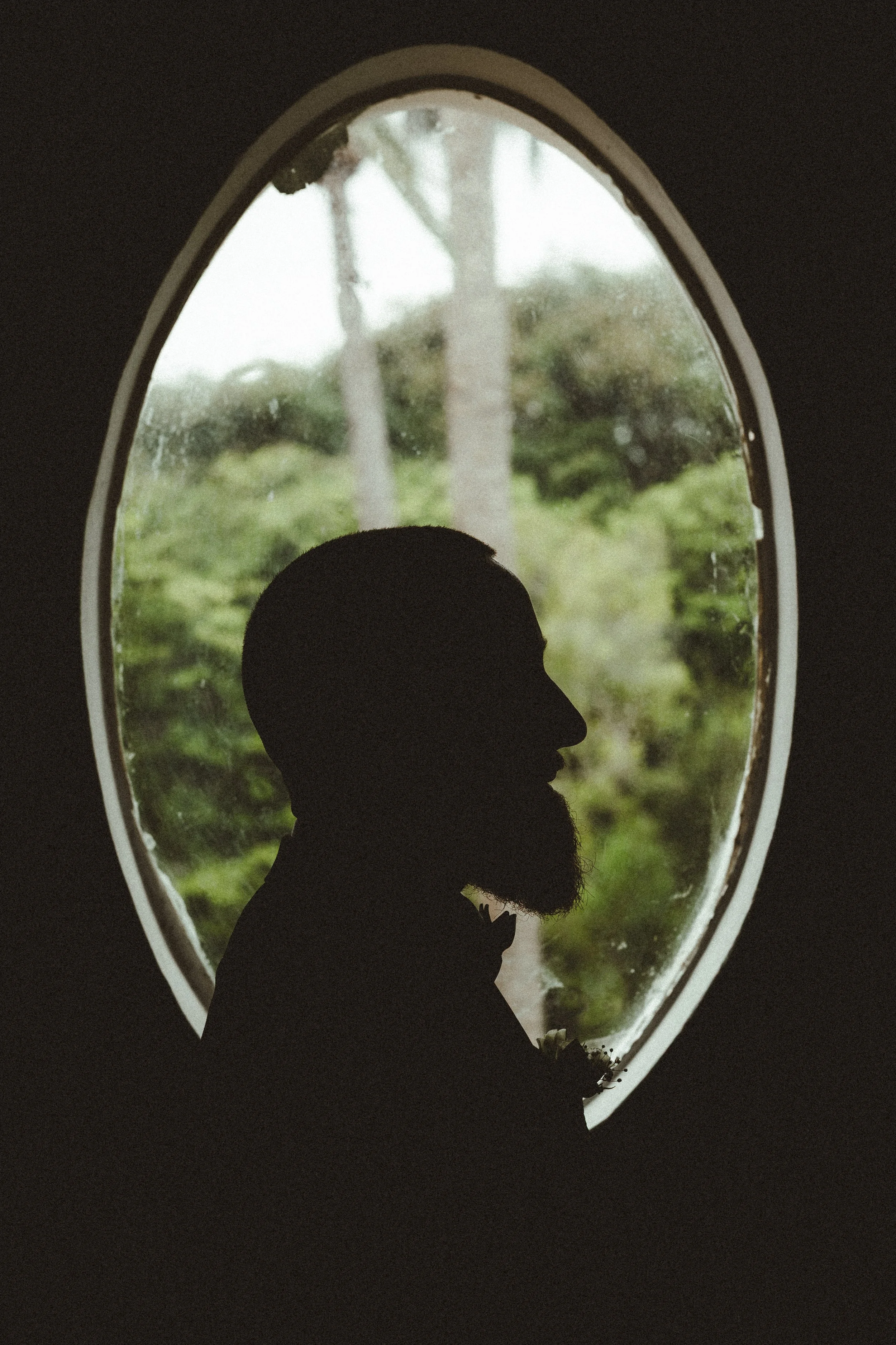 Silhouette of a man with a beard and bow tie in front of an oval window with greenery outside.