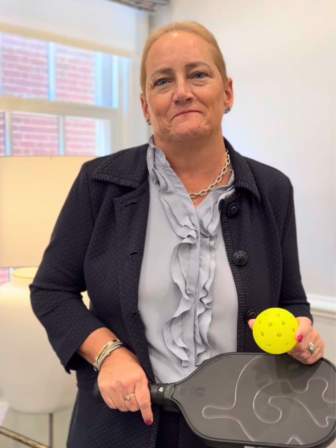 A middle-aged woman with blonde hair stands indoors, holding a yellow ball and a paddle. She wears a dark blazer, a light-colored blouse with ruffles, and a necklace, with a slight smile on her face.