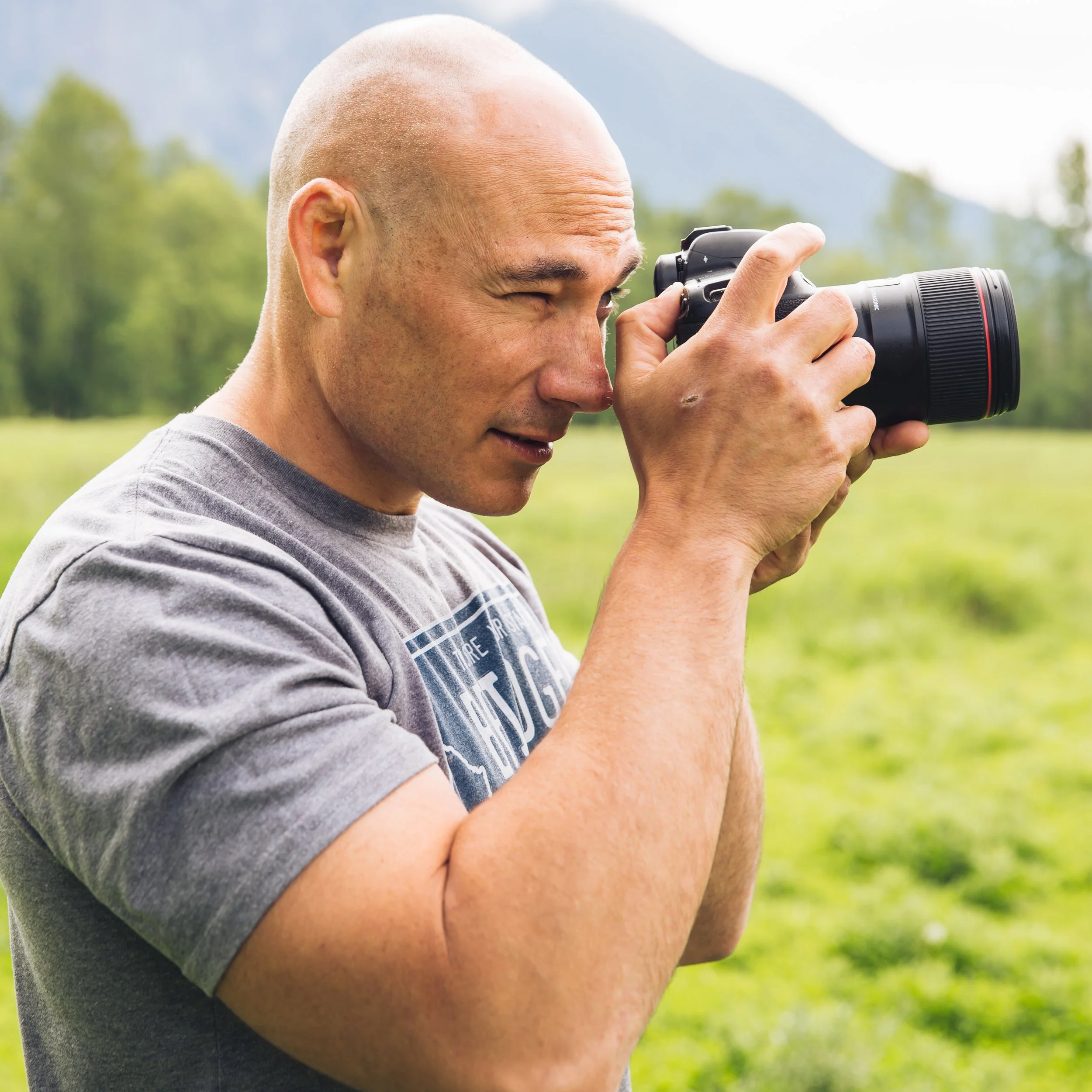 A person with a bald head and tan skin is holding a black camera with a zoom lens, taking a photograph outdoors on a sunny day with a blurred background of trees and mountains.