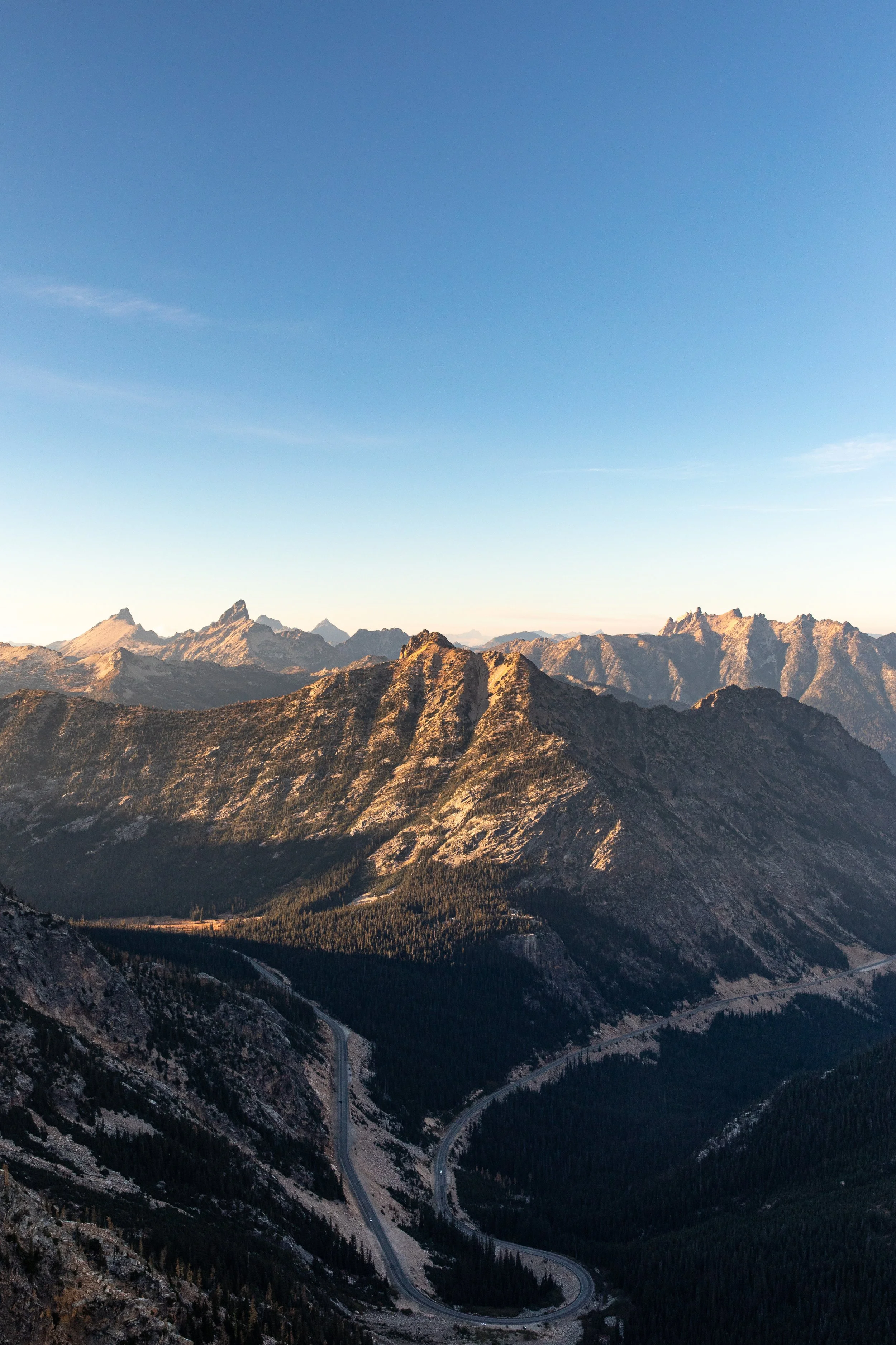 Scenic view of rugged mountain range with rocky peaks, forested slopes, and a winding road at the base, under a clear blue sky.