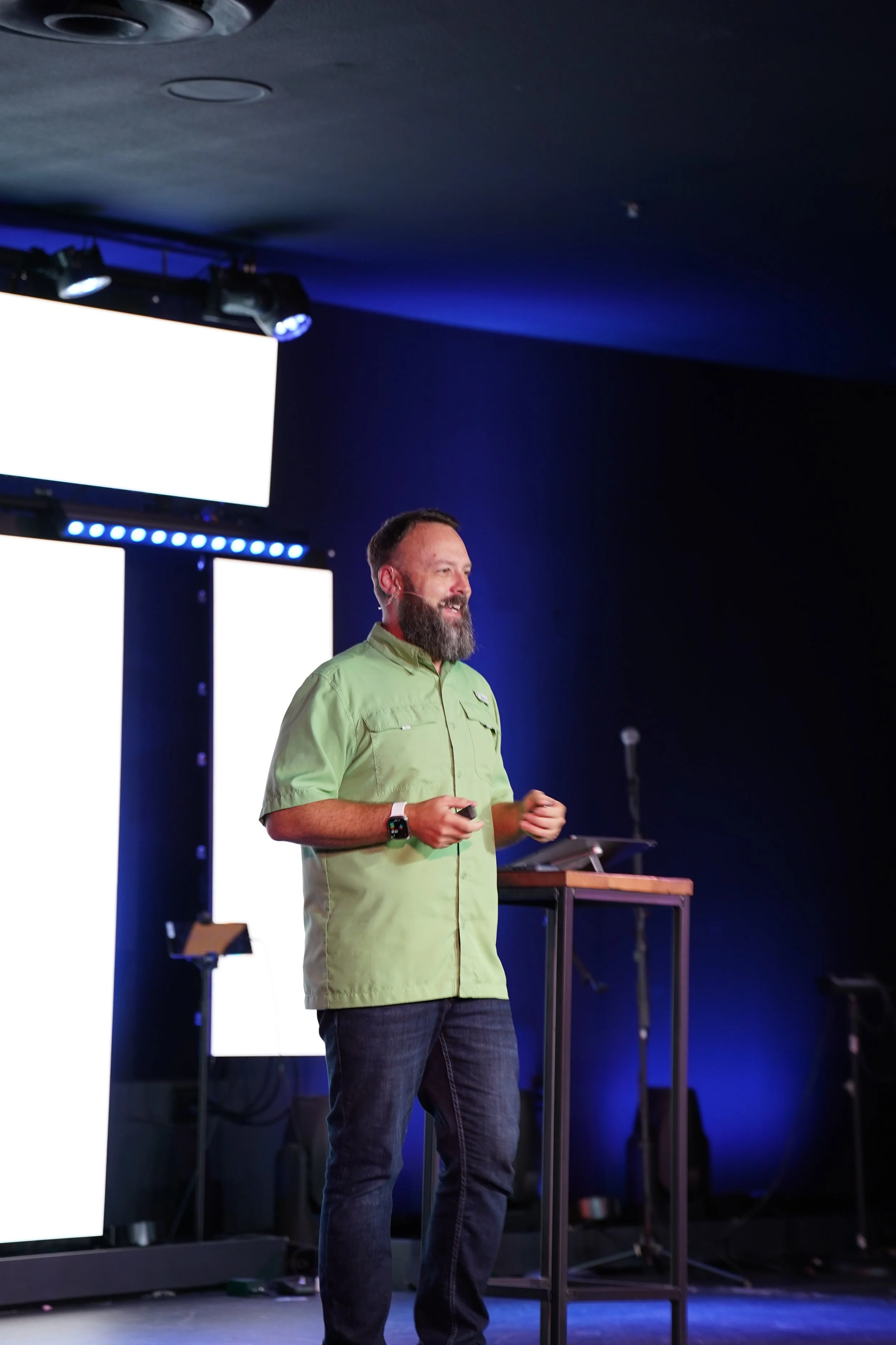 A man with a beard, wearing a white short-sleeved shirt and jeans, stands on a stage holding a microphone and speaking to an audience. There is a yellow chair behind him and purple lighting in the background.