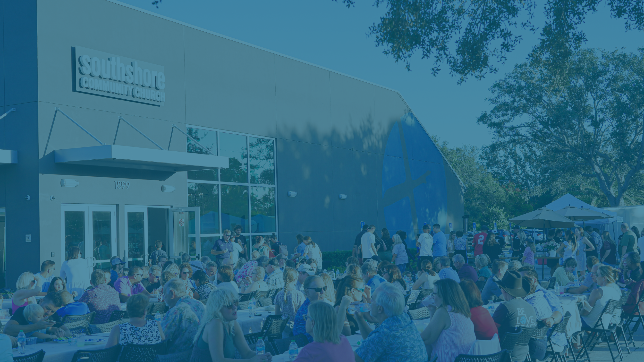 Crowd of people sitting at outdoor tables outside Southshore Community Church during daytime event with tents and trees in background.