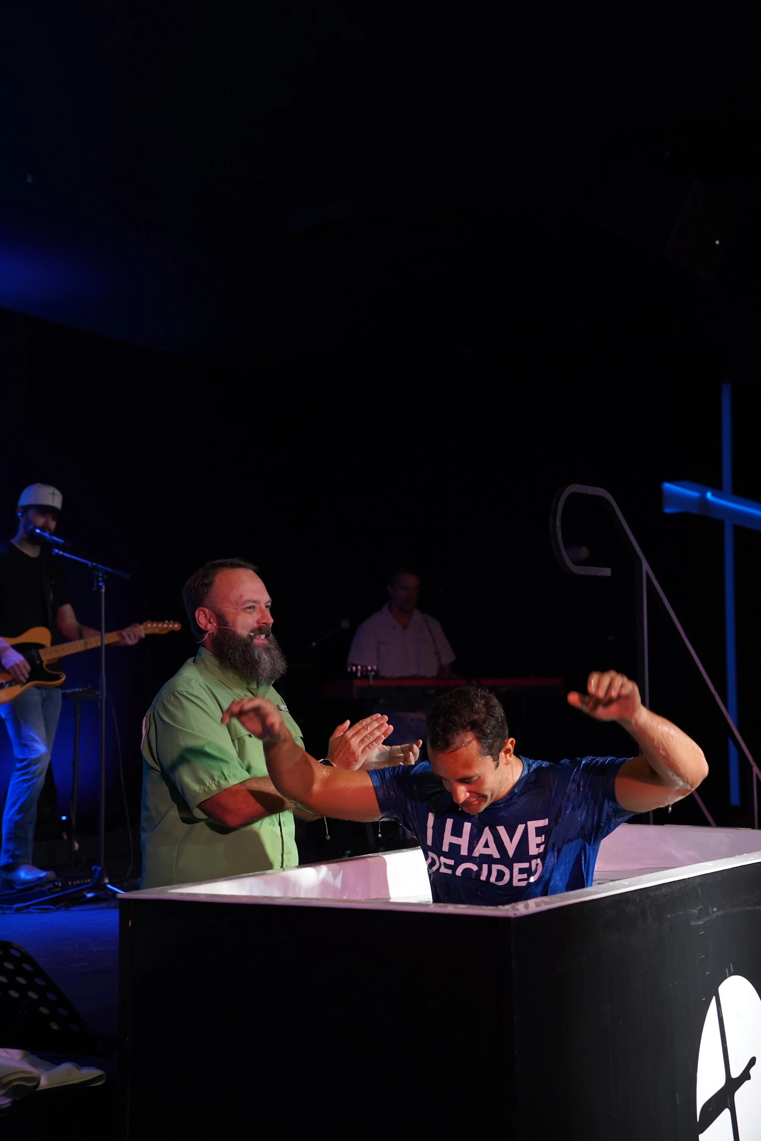 Woman with arms raised in a baptismal pool during a religious ceremony, surrounded by smiling people in a dark room with a blue cross in the background.