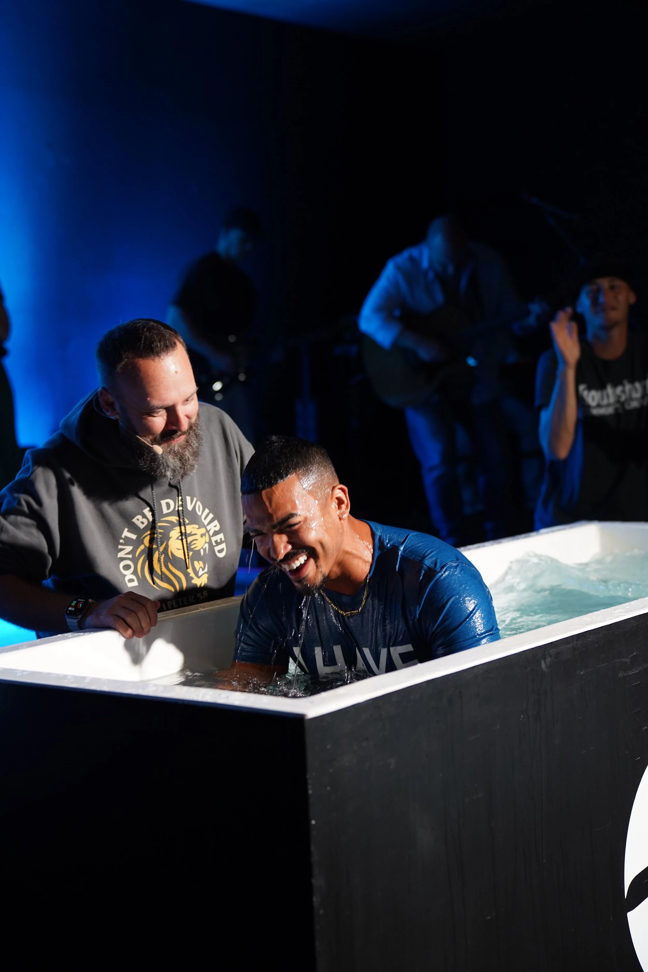 Woman with arms raised in a baptismal pool during a religious ceremony, surrounded by smiling people in a dark room with a blue cross in the background.