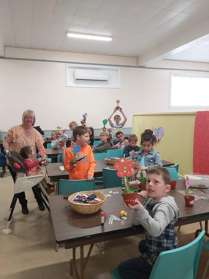 Children in a Sunday School doing arts and crafts, holding decorated flower pots and paper flowers, with a teacher assisting.