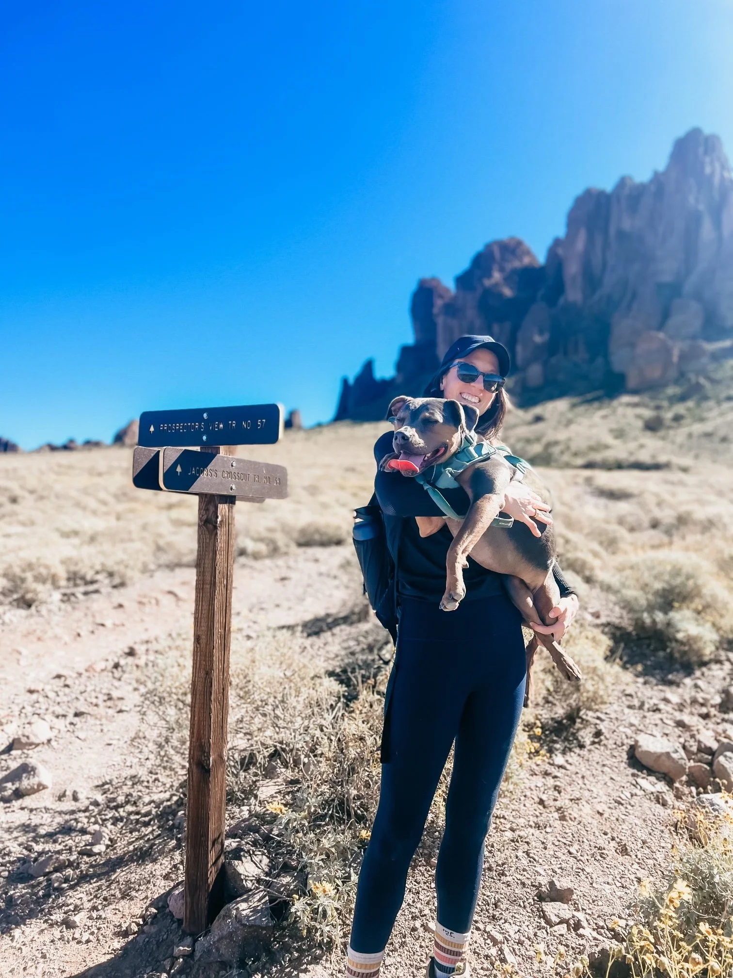 Dr. Elizabeth holding her dog Luna on a desert hiking trail near a trail sign, representing the path of building nervous system capacity and long-term healing.