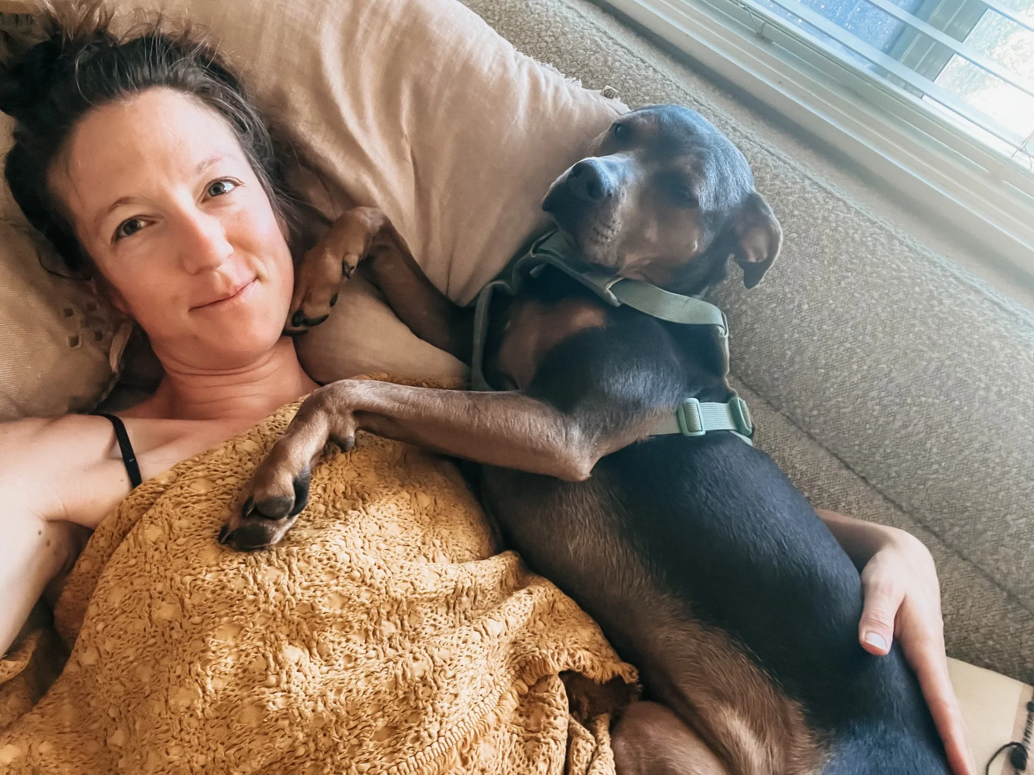 Woman resting on a couch cuddled with her dog, representing nervous system safety, self-trust, and healing through softness rather than self-fixing