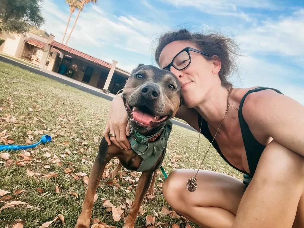 Woman sitting outdoors on grass hugging her smiling dog, reflecting the connection between nervous system safety, emotional regulation, self-trust, and living in alignment.