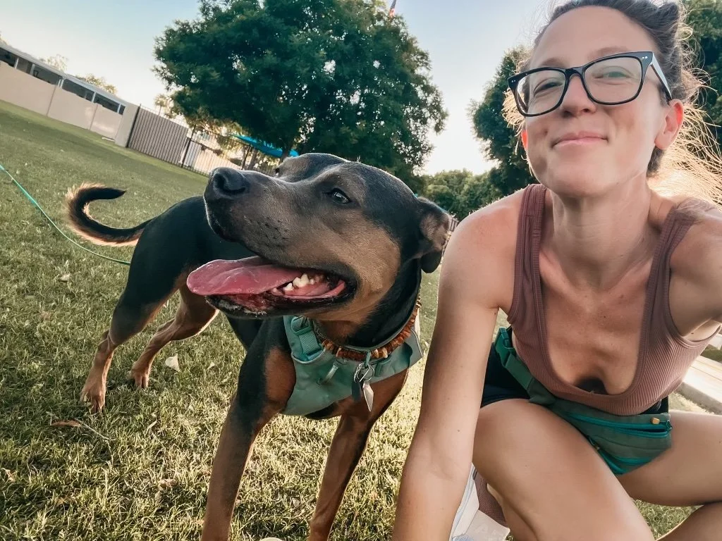 Dr. Elizabeth sitting on the grass outdoors with her two dogs, smiling at the camera during a sunny afternoon.  The dogs sit close beside her, creating a relaxed and joyful moment in a neighborhood park setting.