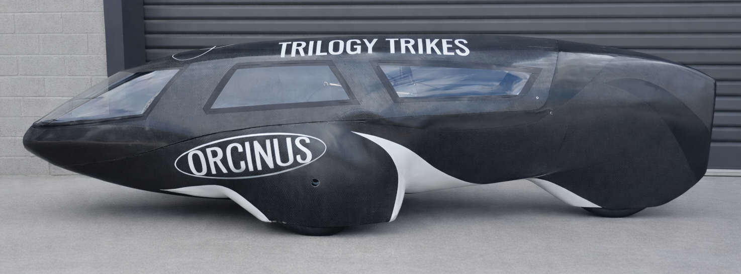 A small black and white racing trike with a streamlined, aerodynamic design and transparent canopy, labeled 'TRILOGY TRIKES' and 'ORCINUS'.