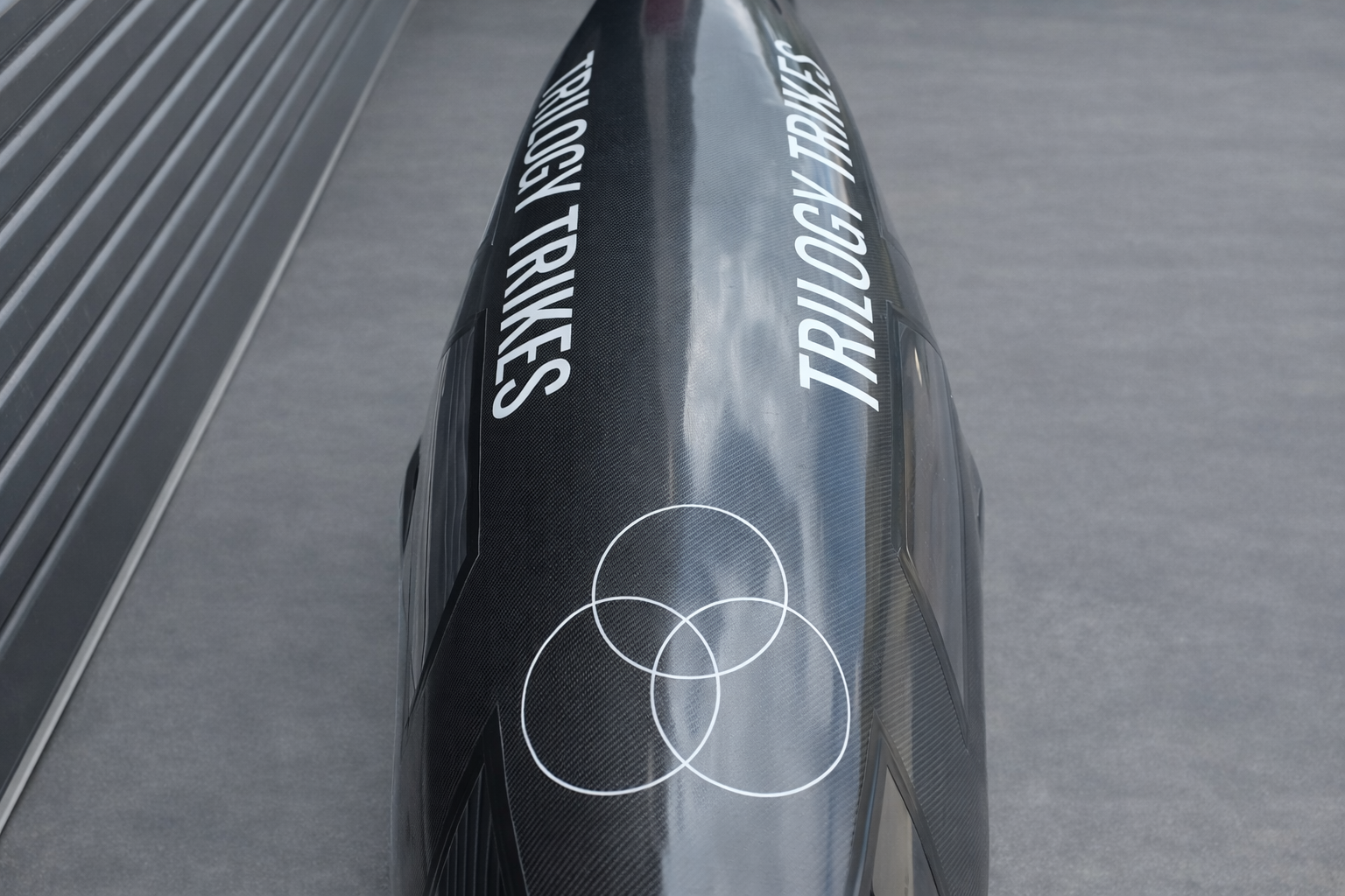 Top view of a black racing trike with white text and a logo, showing the carbon fibre pattern.