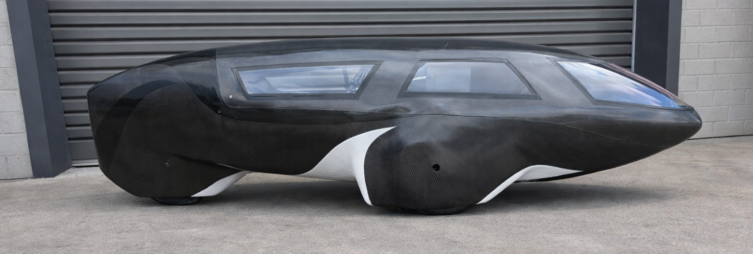 A futuristic, black and white trike with a sleek, aerodynamic design, shown parked outside near a gray brick wall and a metal shutter.