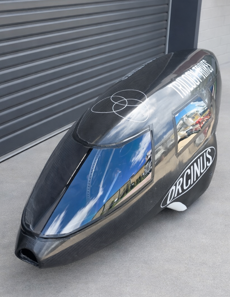 A black carbon fiber streamlined racing trike with a large windshield, parked on a concrete surface near a gray metal wall, with the word "ORCID" on its side and a logo on the top.
