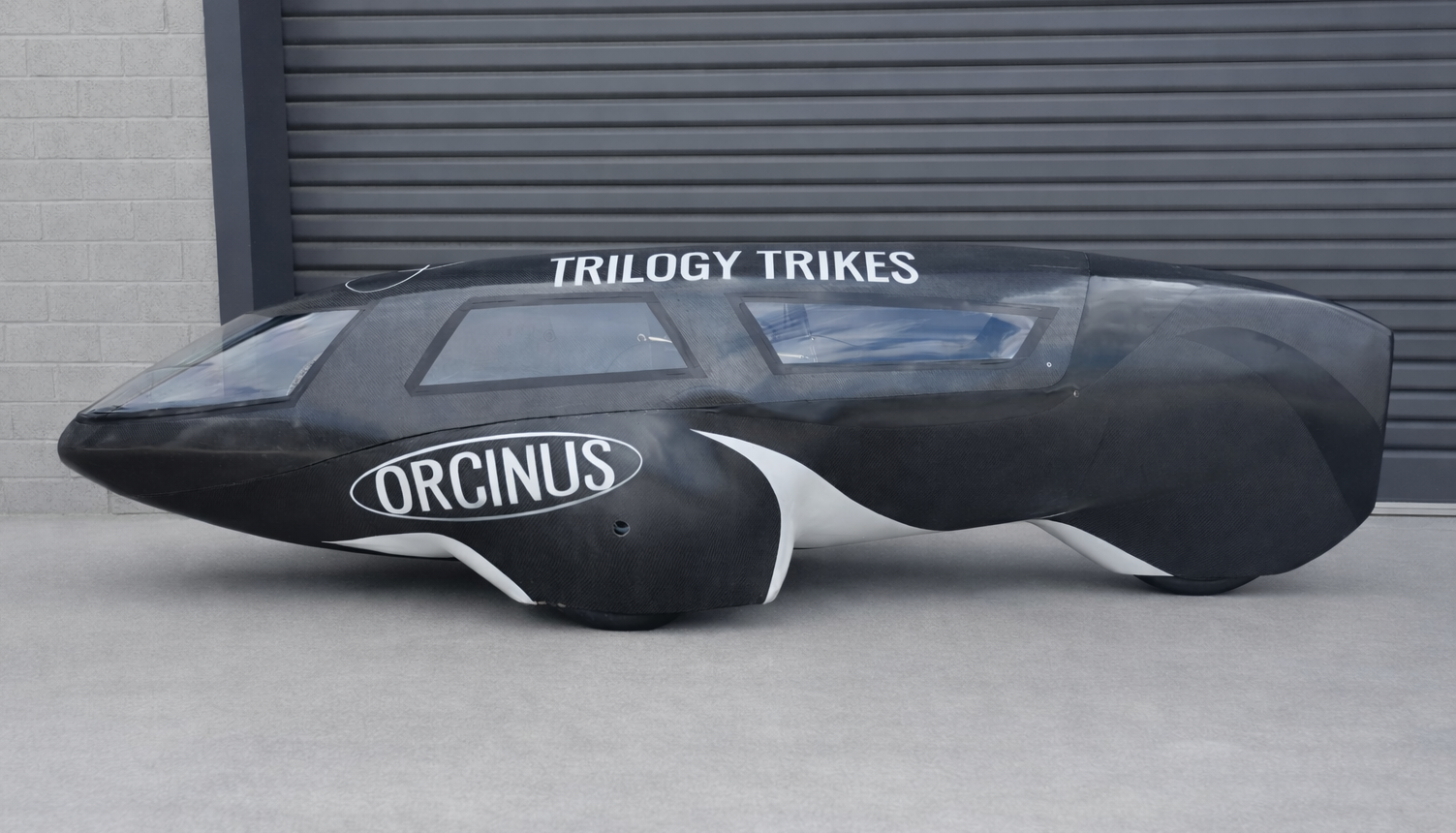 Black race trike with sleek aerodynamic design, labeled with 'Orcinus' and 'Trilogy Trikes' on the sides, parked on concrete ground in front of a gray garage door.