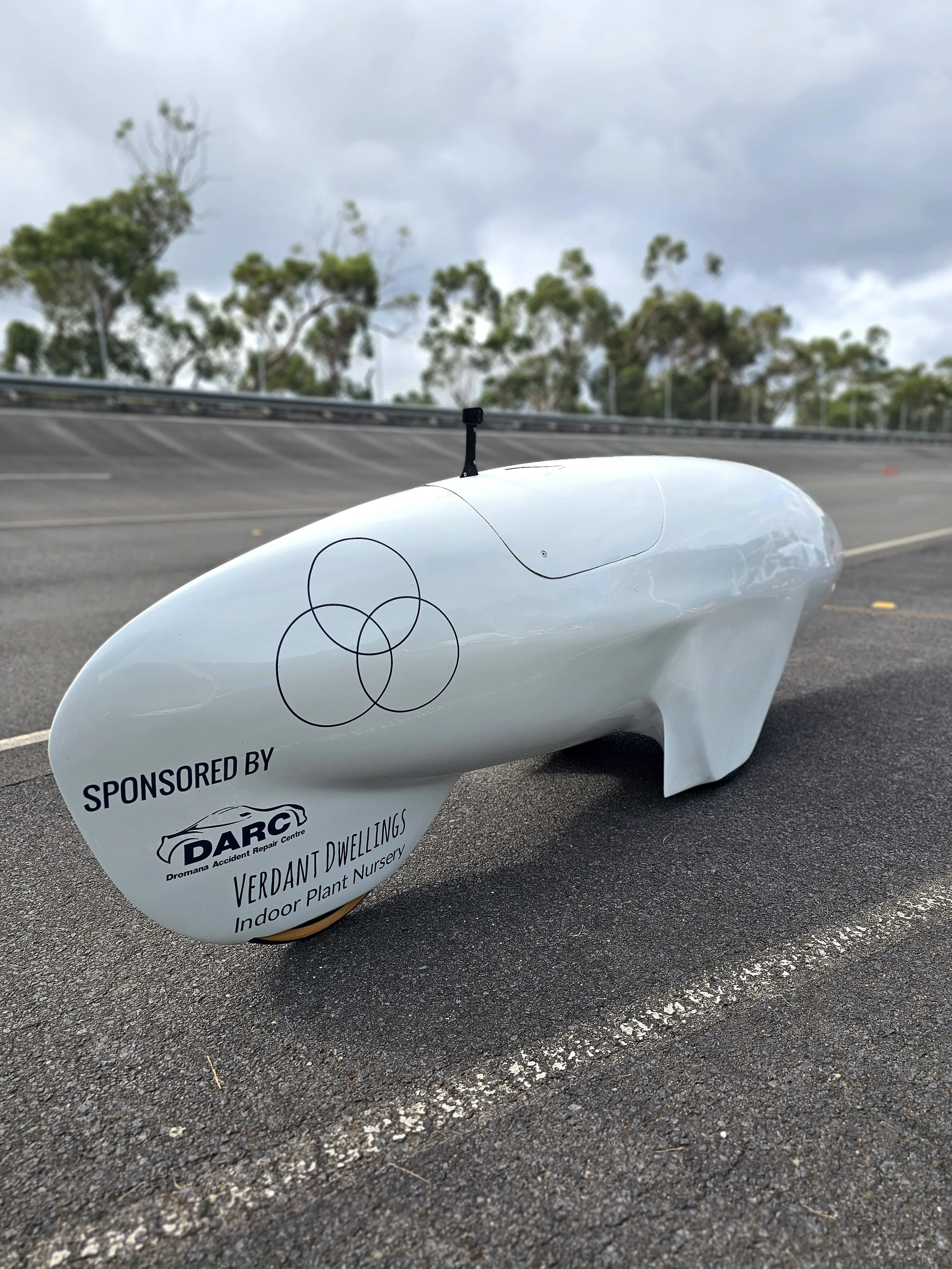 A sleek, modern trike, parked on an asphalt surface in an outdoor area with trees and cloudy sky in the background. The vehicle is sponsored by Verdant Dwellings and Dromana Accident Repair Centre, with a logo and text on its side.