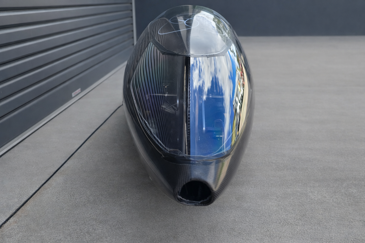 A sleek, black, futuristic trike with a transparent canopy, reflecting a cloudy sky and surrounding environment, parked on a gray surface near a corrugated metal wall.
