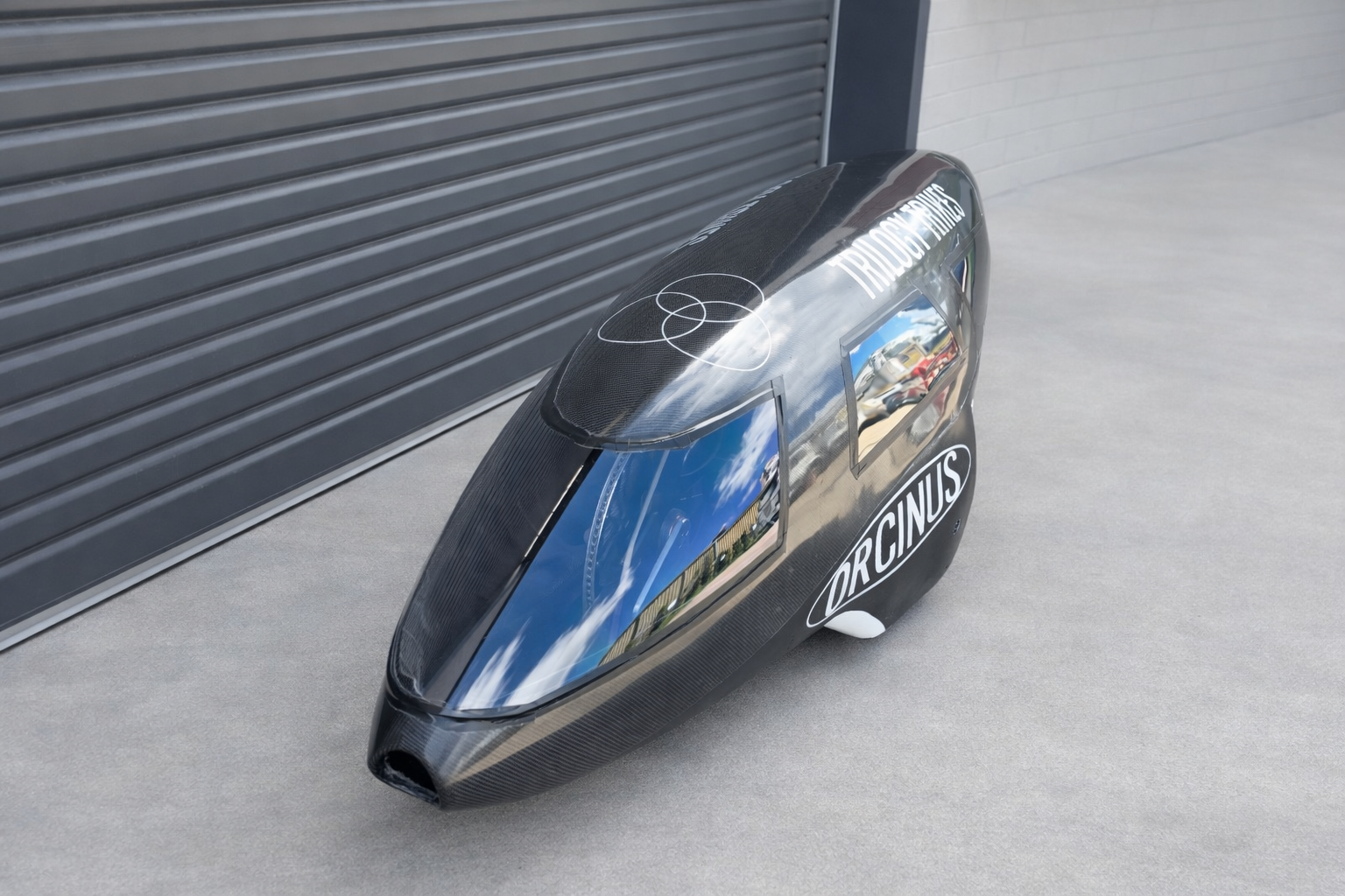 A sleek, black trike with a clear canopy, labelled "ORCINUS" on the side, parked outside near a steel roller door.