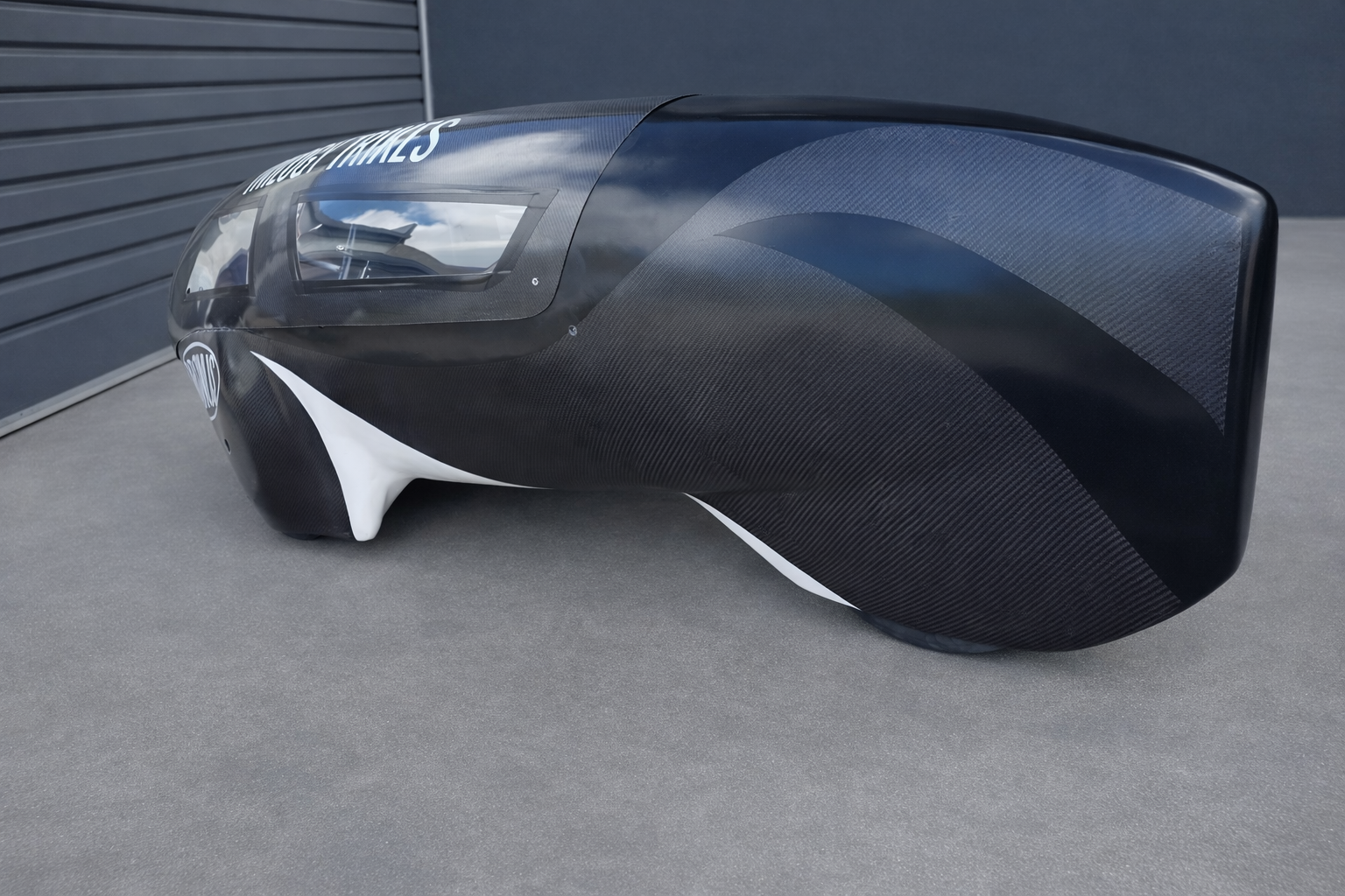 A sleek, black and white racing trike with a low profile and aerodynamic design, placed on a gray surface against a gray wall background.