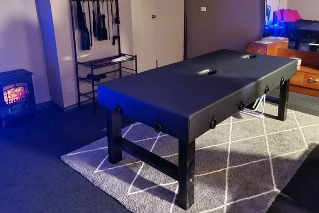 Custom black milking table in a dimly lit Melbourne massage room with bondage gear, restraints, full-length mirror, and cosy electric fireplace ambiance.