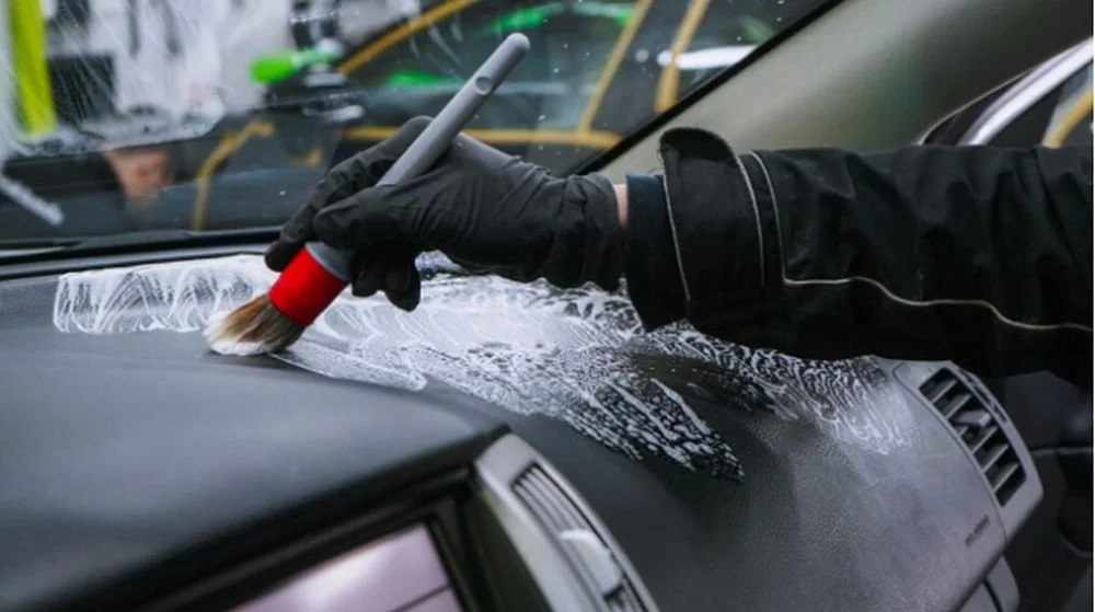 Person wearing black gloves applying cleaning foam to a car dashboard with a brush