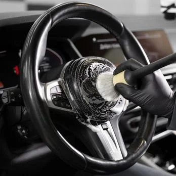 A person wearing a black glove cleaning a car steering wheel with a brush and cleaning foam.