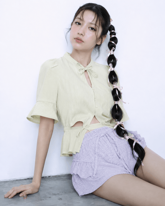 Young woman with long hair styled in large braids with ribbons, wearing a light yellow blouse and lavender shorts, sitting against a white wall.